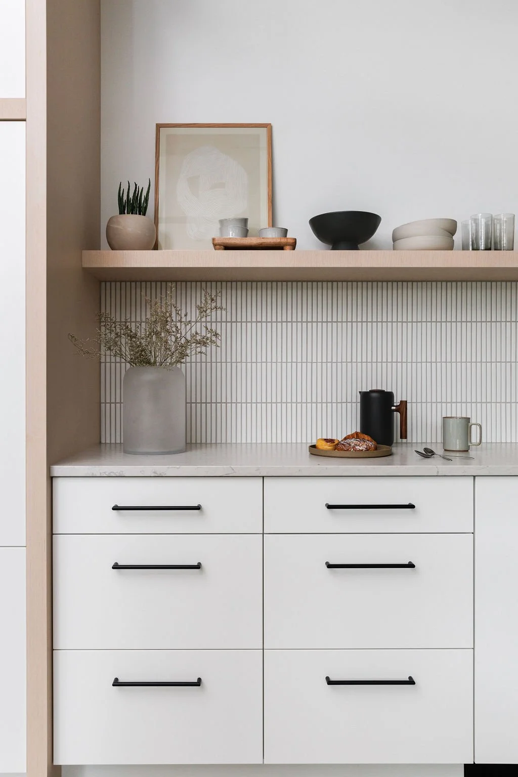 White kitchen with black accents, floating shelf with art and decor and small rectangle white mosaic tiles.