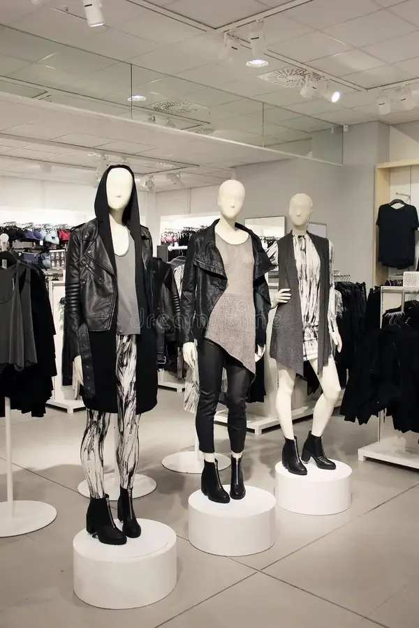 Three mannequins in a clothing store display wearing black and gray outfits. The first mannequin is dressed in a black leather jacket, a gray top, patterned leggings, and black boots. The second mannequin wears a black leather jacket over a gray dres