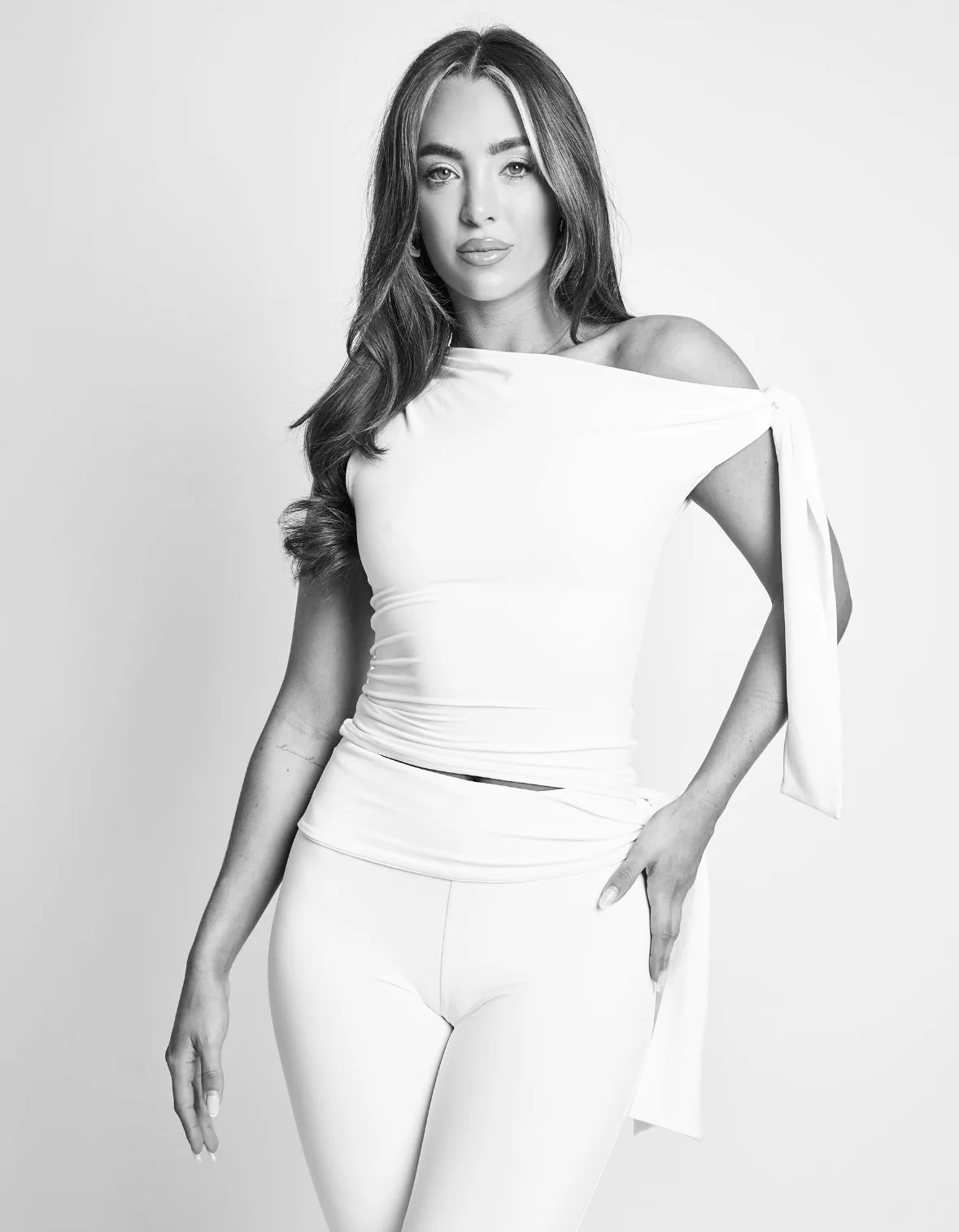 Black and white portrait of a woman with long wavy hair, wearing an off-shoulder top with a tied sleeve and fitted pants, posing against a plain background.
