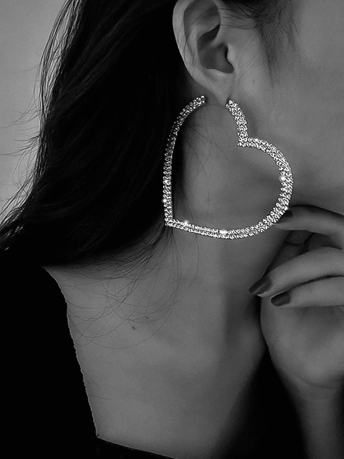 Close-up of a woman's face and neck, wearing a large, heart-shaped earring decorated with small sparkling stones, with her hand resting near her chin.