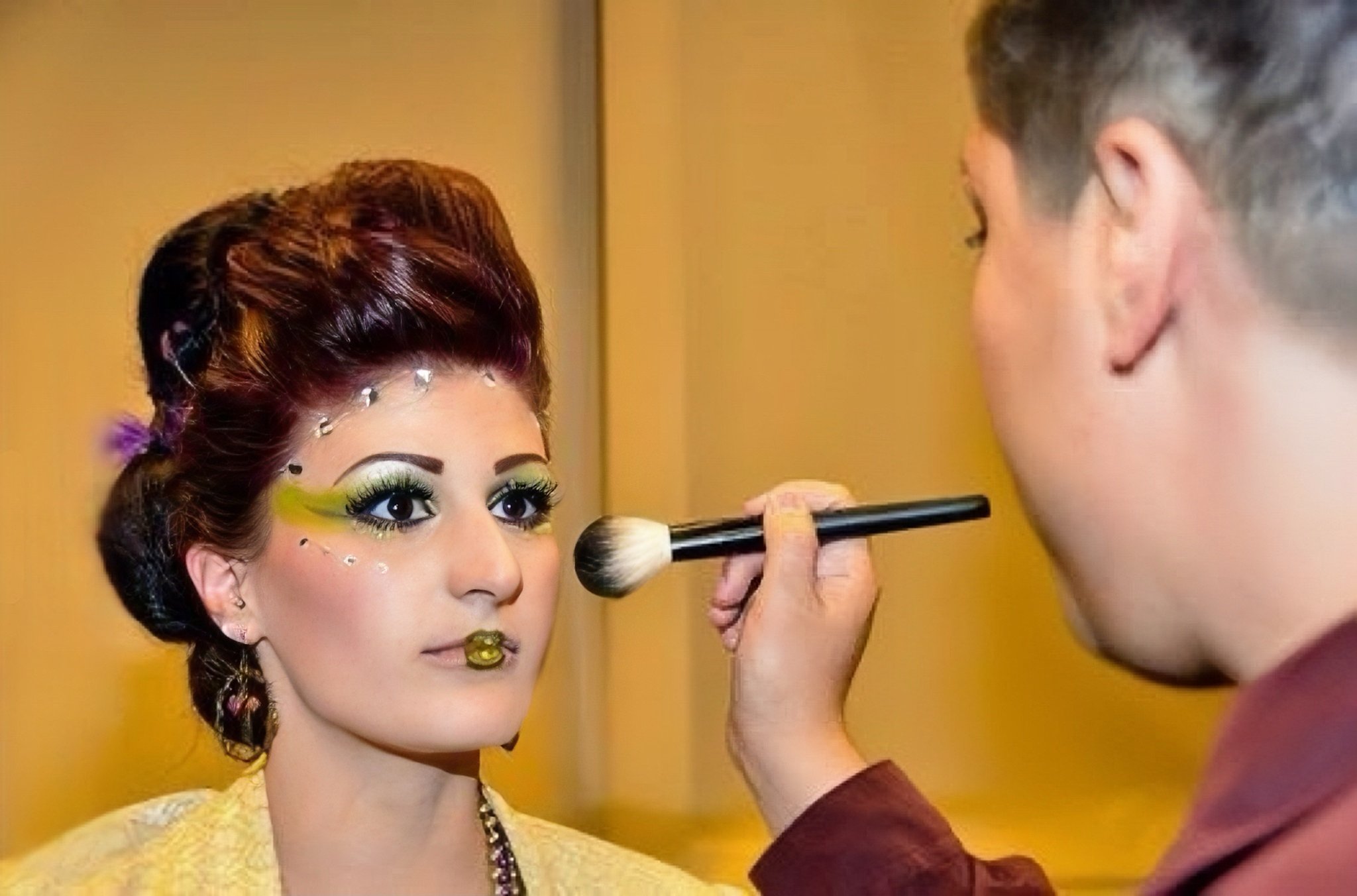 A woman getting her makeup done by a makeup artist using a large makeup brush, with vibrant eye makeup, jewelry, and her hair styled elaborately.