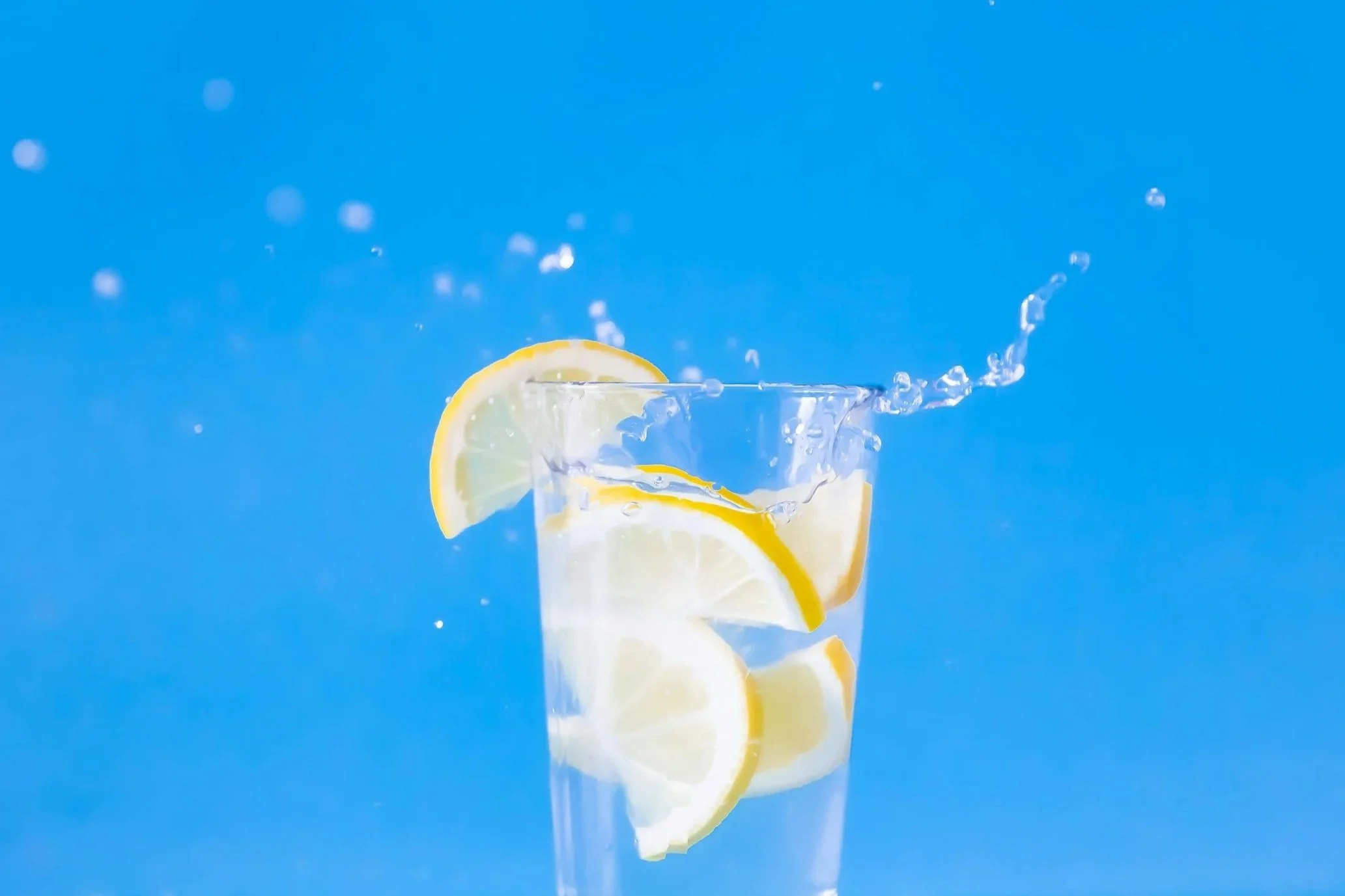 A clear glass filled with water and lemon slices, with a slice of lemon on the rim. Water splashes out as the glass is shaken or dropped, set against a bright blue background.