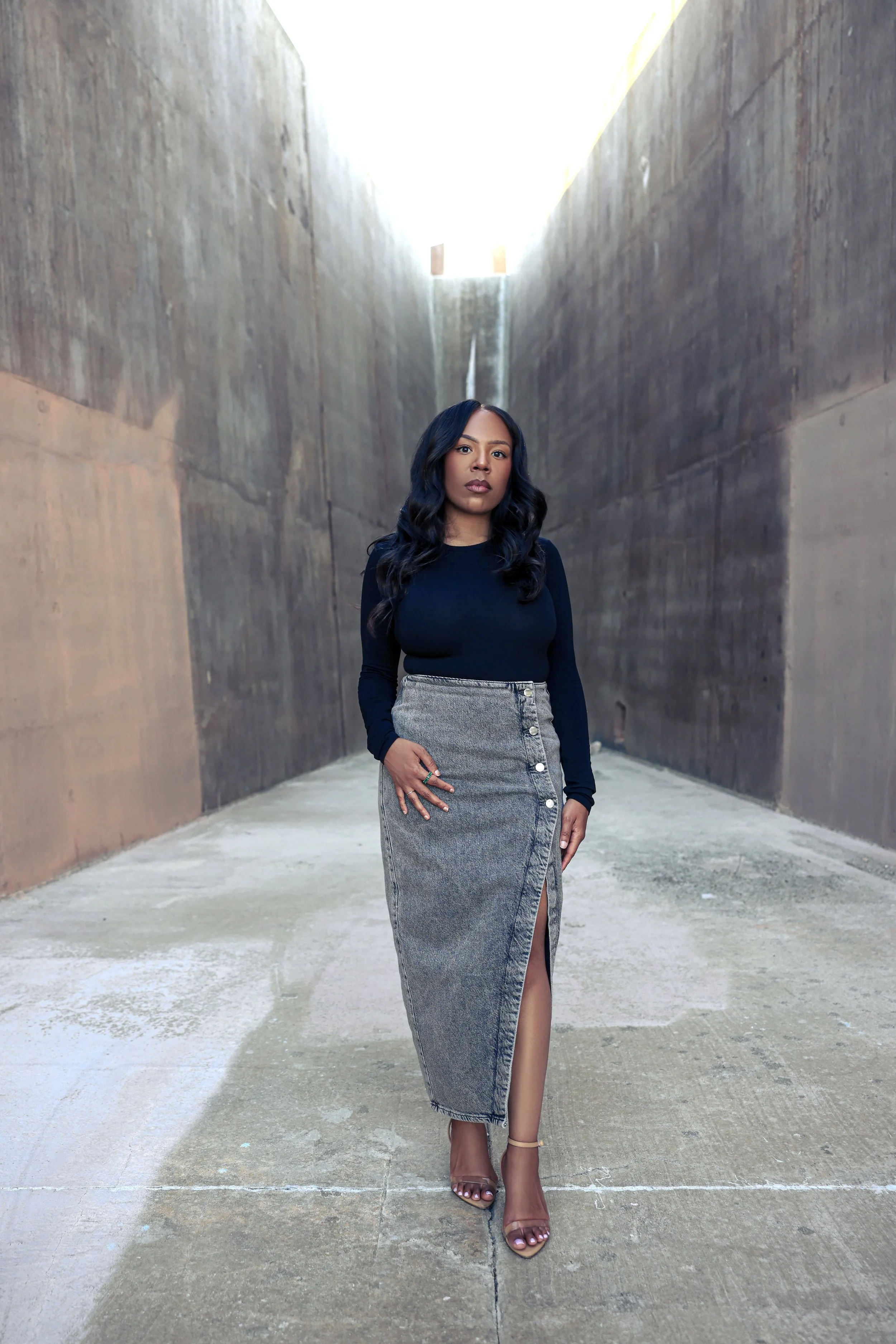 A woman with long dark wavy hair wearing a black long-sleeve top and a high-slit gray denim skirt standing in an empty, narrow concrete alleyway with high walls. The sky is bright above, and she is wearing transparent high-heeled sandals.