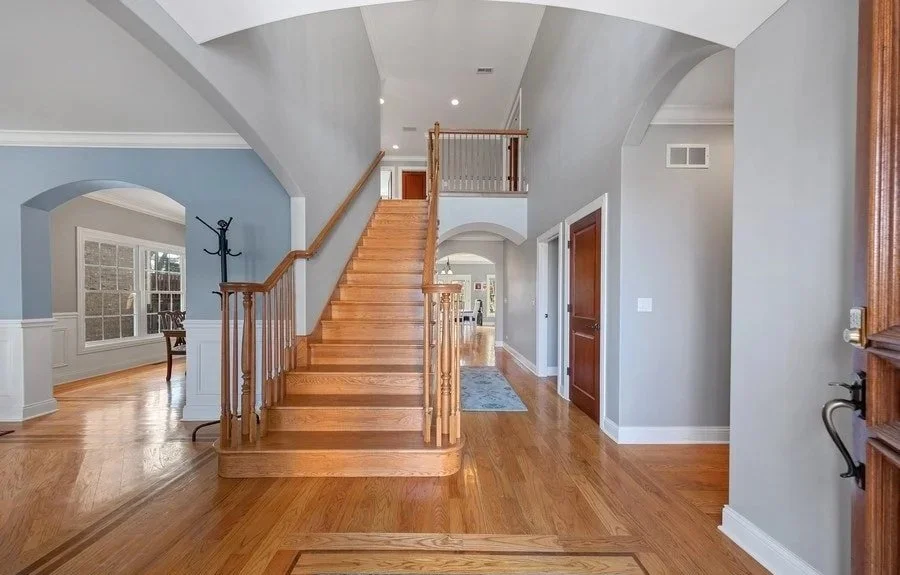 Interior of a house with a wooden staircase leading to the second floor, hardwood floors, light gray walls, and trim. There is a hallway and a view into the dining room and living area.