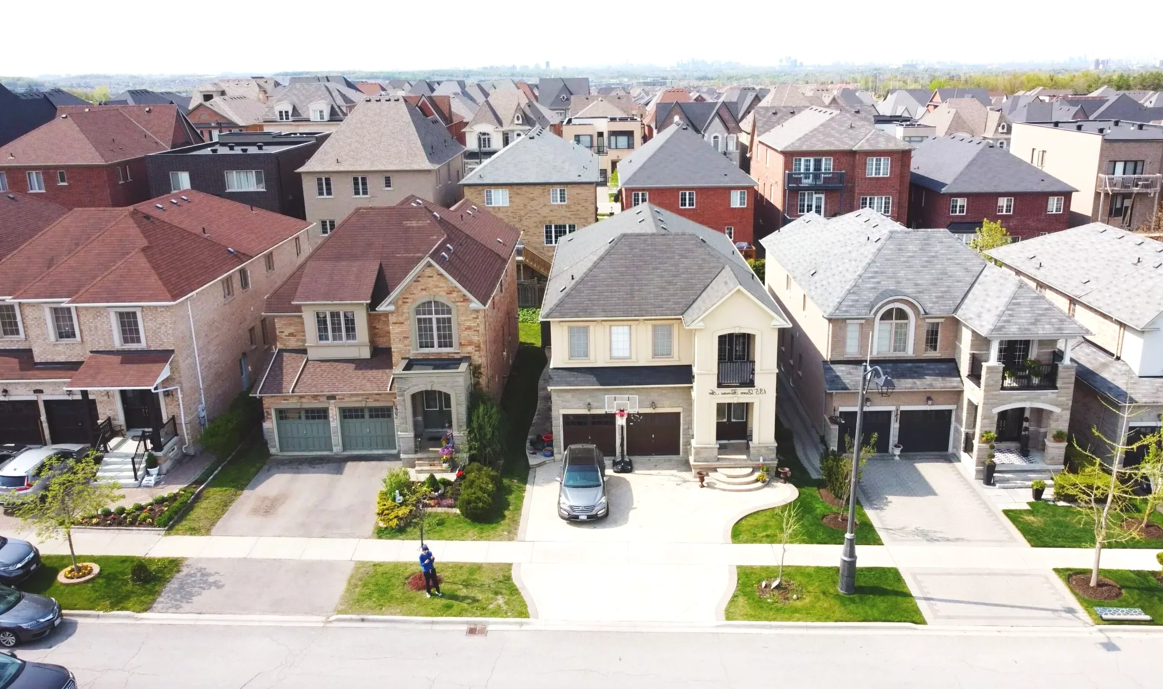 A row of suburban houses with driveways, front yards, and street lined with trees and lampposts.