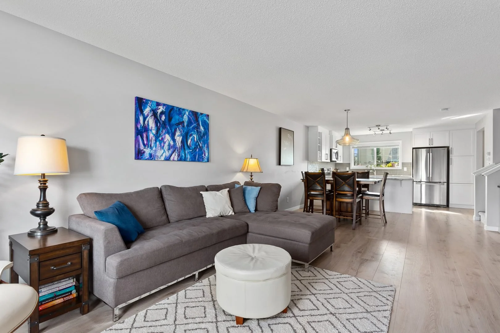 Open-concept living room and kitchen with gray sectional sofa, a white round ottoman, wooden side table, and artwork on the wall, in a modern home.
