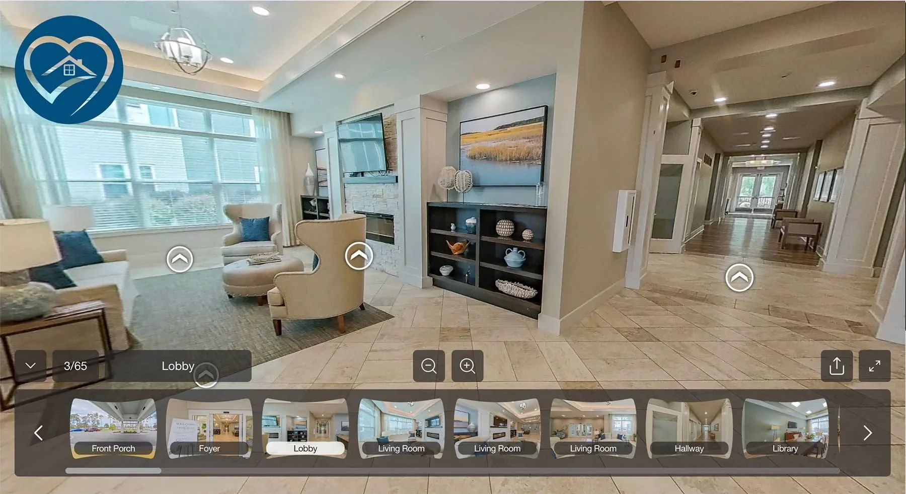 Interior view of a spacious, well-lit home lobby area with large window, white couches, an armchair, and a black shelf with decorative items. There is a hallway leading to other rooms, with wooden floors and framed artwork on the walls.