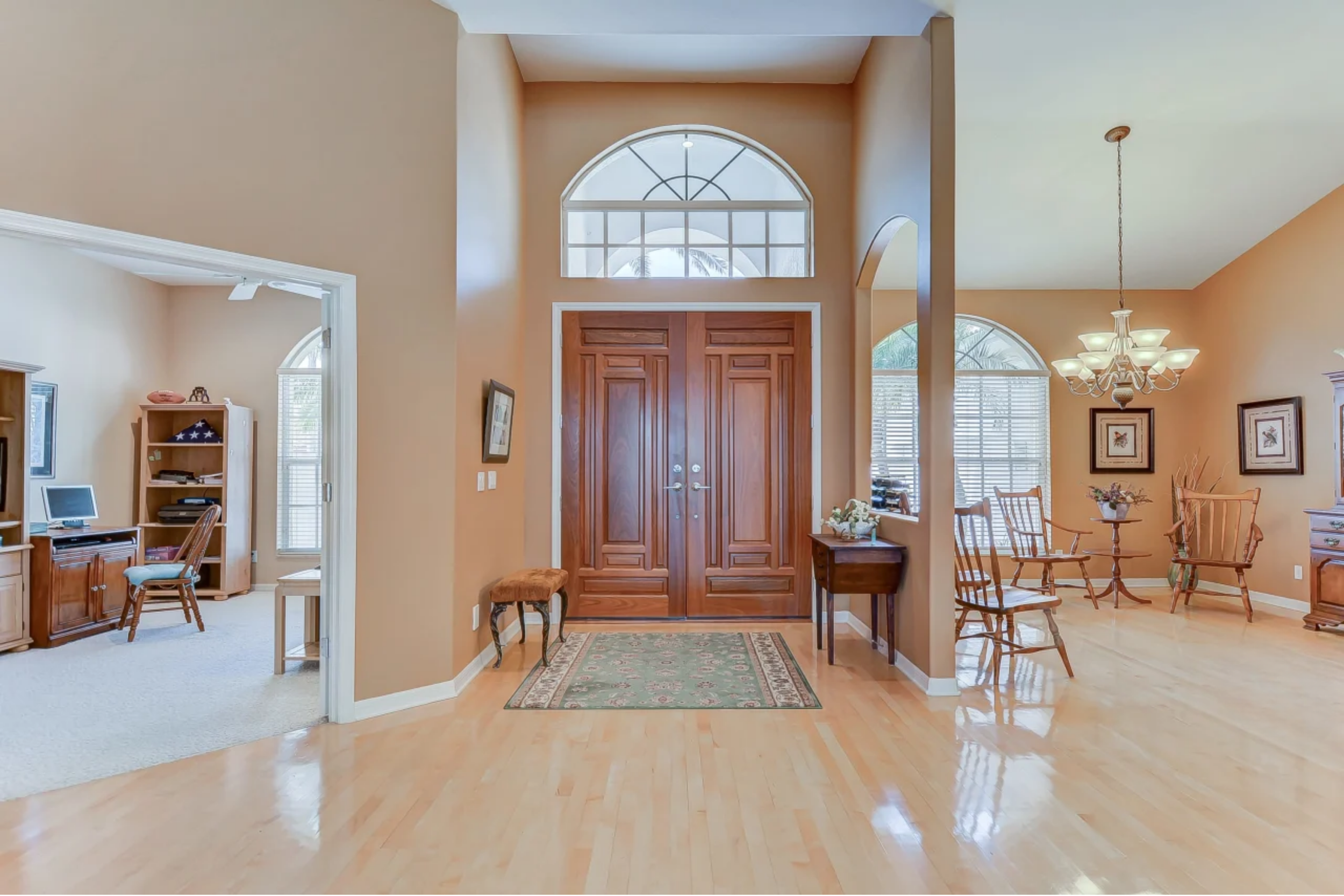 Interior view of a house entryway with a double wooden door, a semi-circular window above, a small rug on the wooden floor, and seats with a side table. To the right, a dining area with wooden chairs, a chandelier, and framed pictures on the wall. To