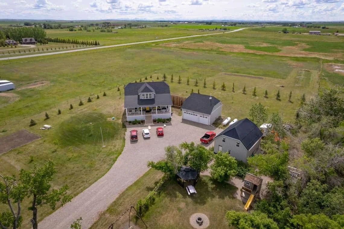 Aerial view of a rural property with a main house, garage, and outbuildings surrounded by open fields and trees.