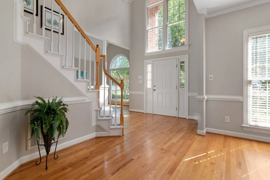 The image shows a bright, spacious living area with hardwood flooring, large windows, and a white front door. There is a staircase with white risers and wooden handrails on the left side, and a potted fern plant is placed at the bottom of the stairs.