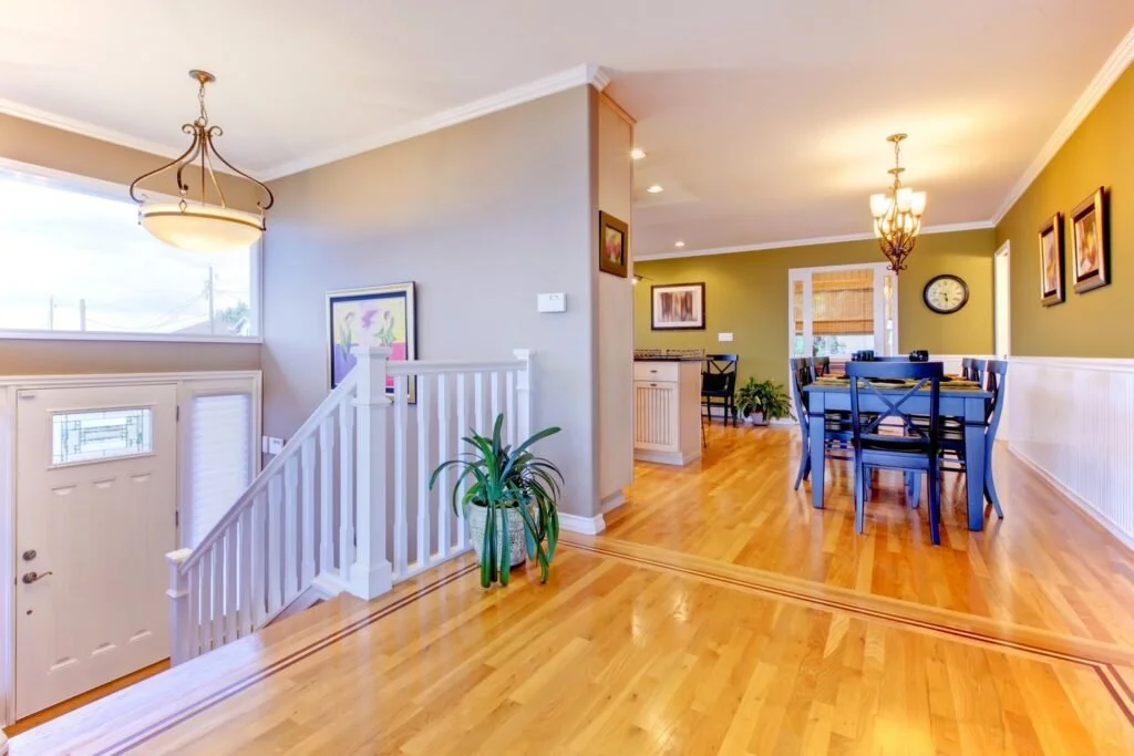 Bright dining area with blue dining set, green wall, chandelier, and framed art, adjacent to kitchen with hardwood floors and staircase with plant