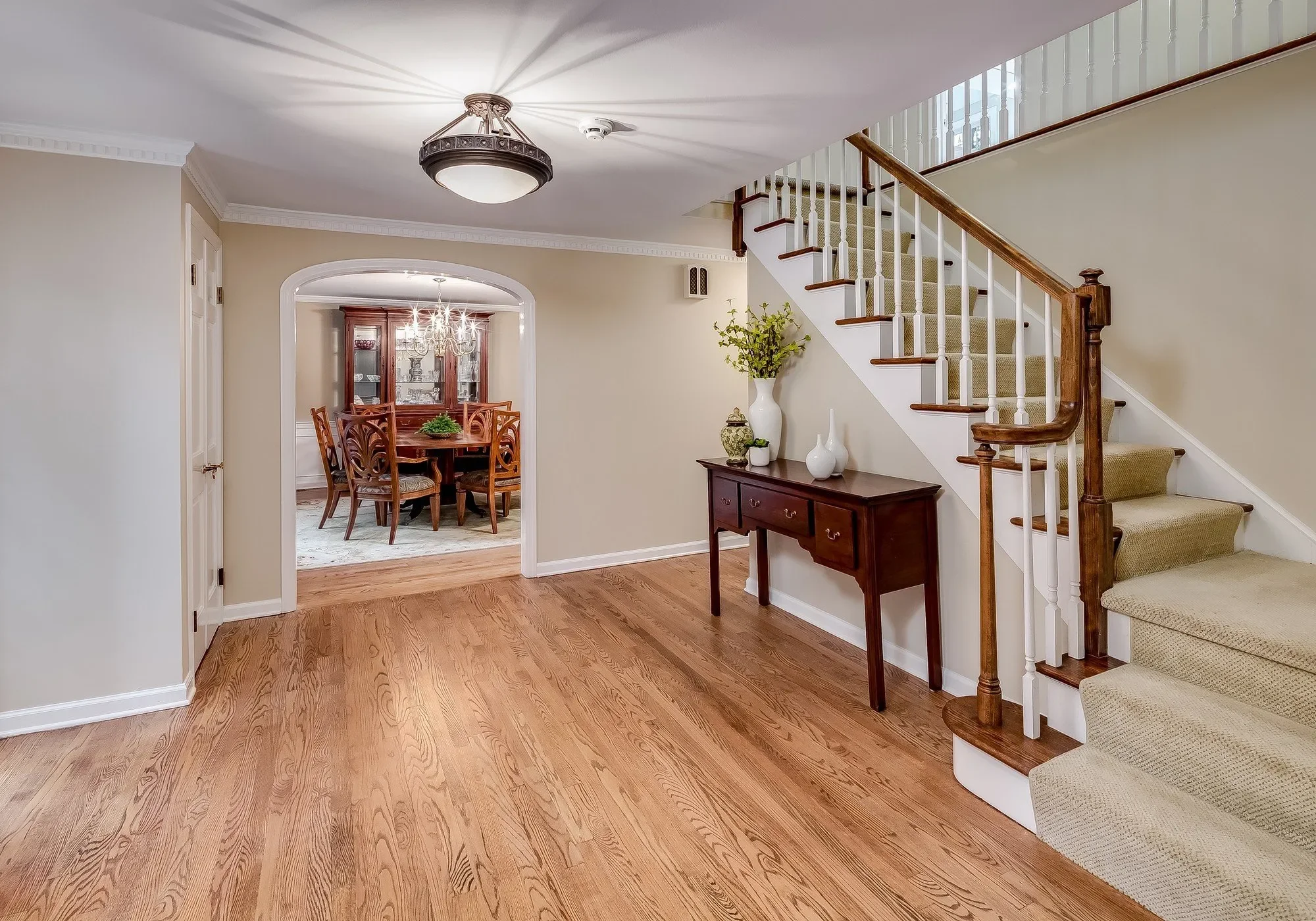Living room with hardwood floors, staircase with beige carpeting, a dark wood console table with white vases, and a doorway leading to a dining room with a round table and wooden chairs.