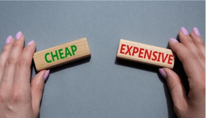 Two hands holding wooden blocks, one with the word 'CHEAP' in green and the other with the word 'EXPENSIVE' in red, separated on a gray surface.