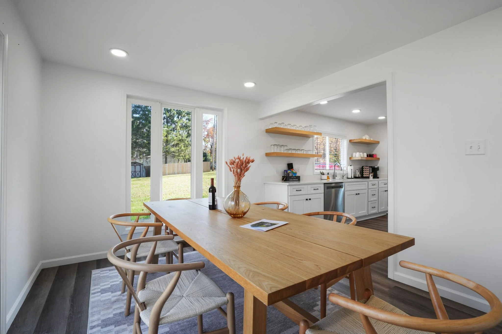 Dining area with a wooden table and six chairs, a vase with pink dried flowers, and a bottle of wine. Open kitchen in the background with white cabinets, open shelves with glassware, a window, and a sliding glass door leading outside to a backyard.
