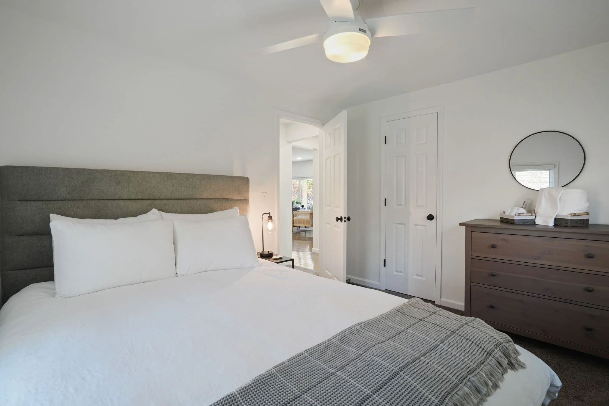 Bedroom with a bed, pillows, bedside table with lamp, dresser with mirror, and open door leading to living area.