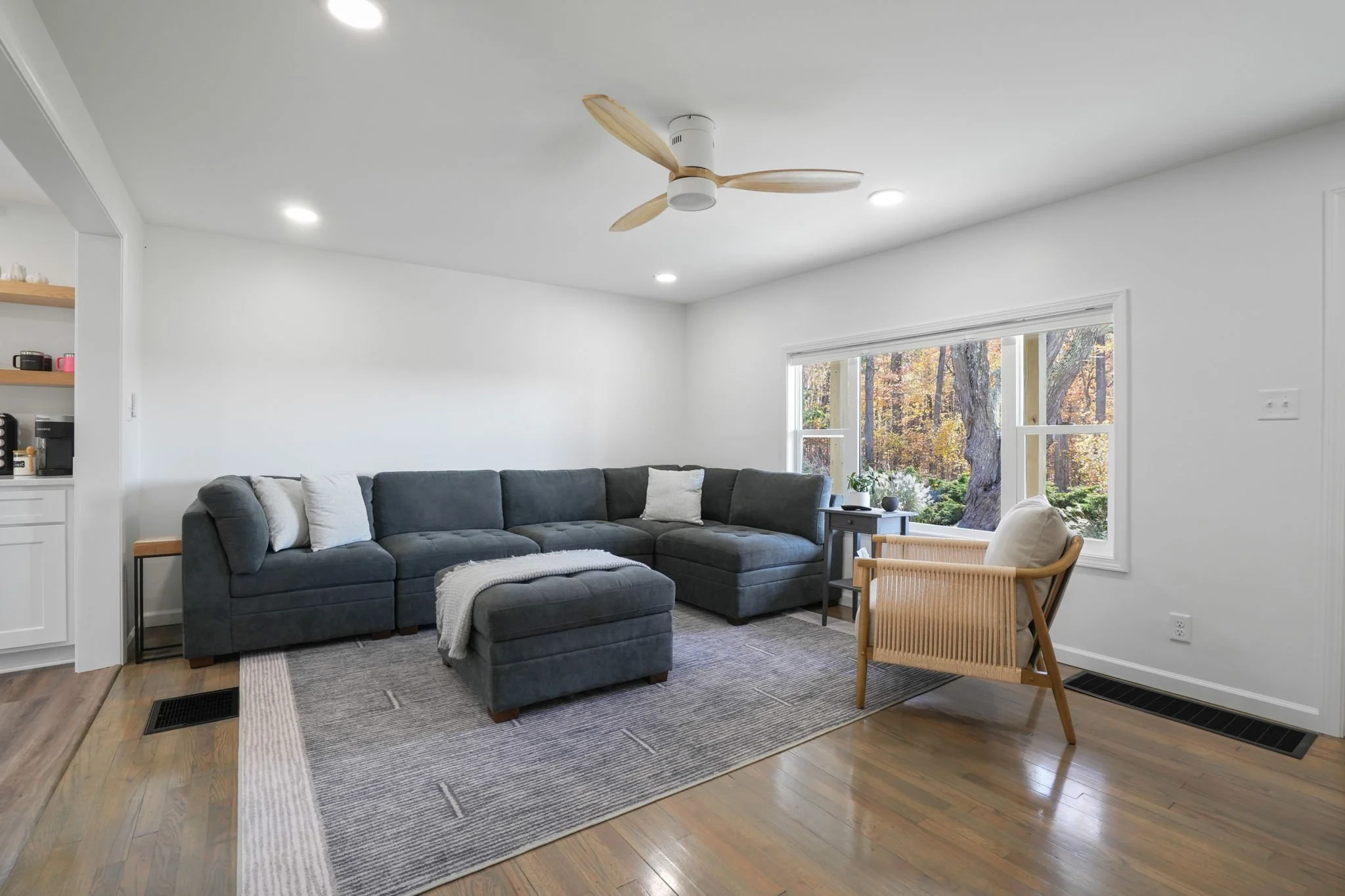 Living room with a large dark gray sectional sofa, a beige armchair, a patterned rug, large window with view of trees, white walls, and wood flooring.