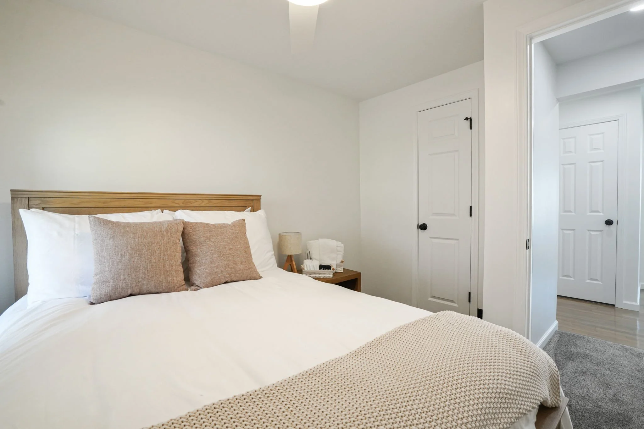A neatly made bed with white linens and beige pillows in a bright, minimalistic bedroom. A small wooden nightstand with a lamp and some towels is beside the bed. Two white doors are visible, one closed and one open leading to another room.