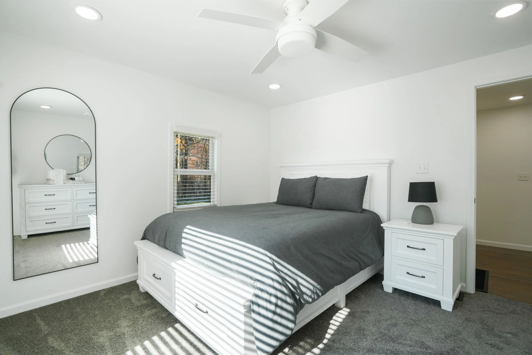 A bedroom with white walls and ceiling, a white bed with gray bedding, a white nightstand with a black lamp, a window with blinds, a ceiling fan, and a large mirror reflecting part of the room.