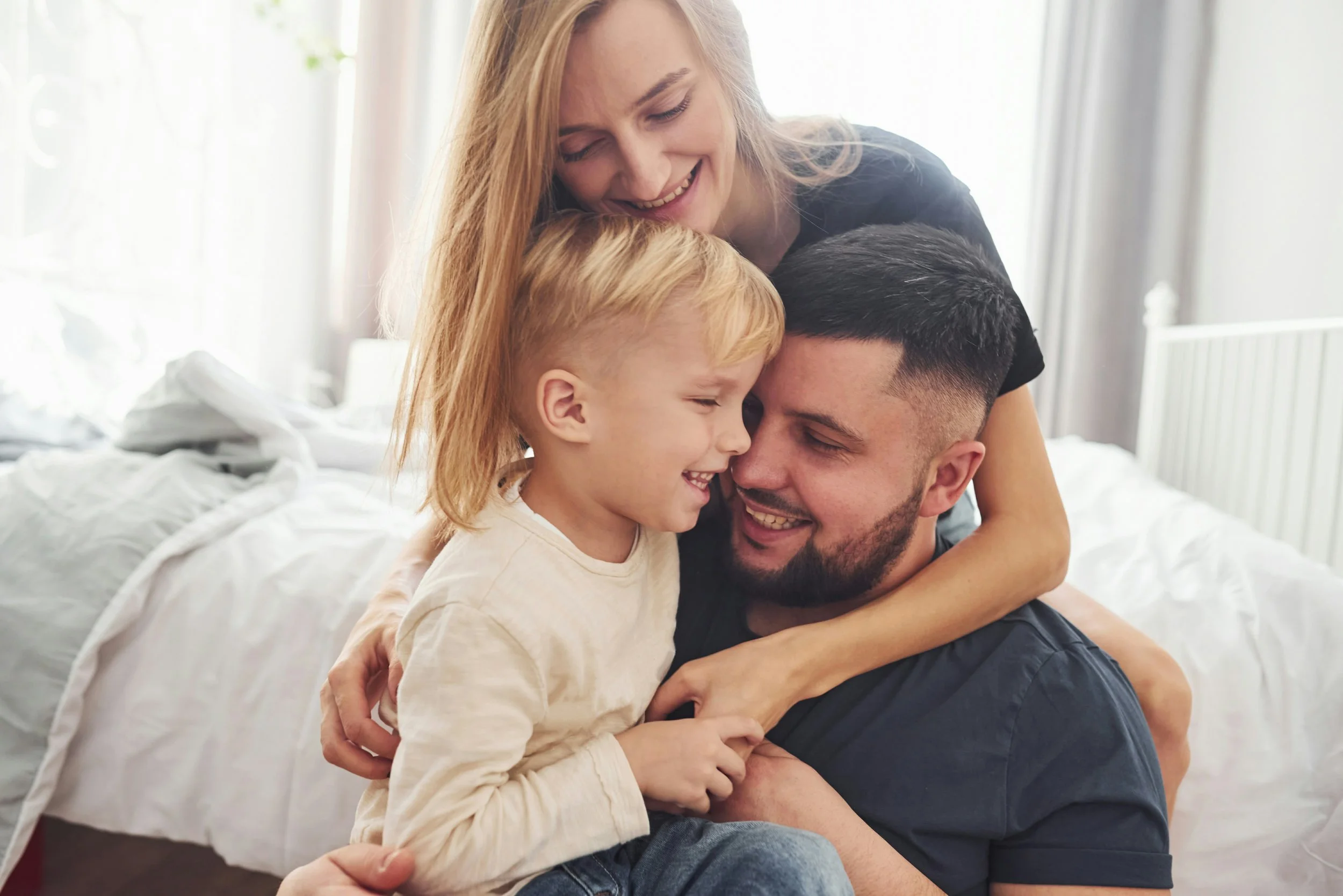 A happy family of four hugging and smiling in a bright bedroom.