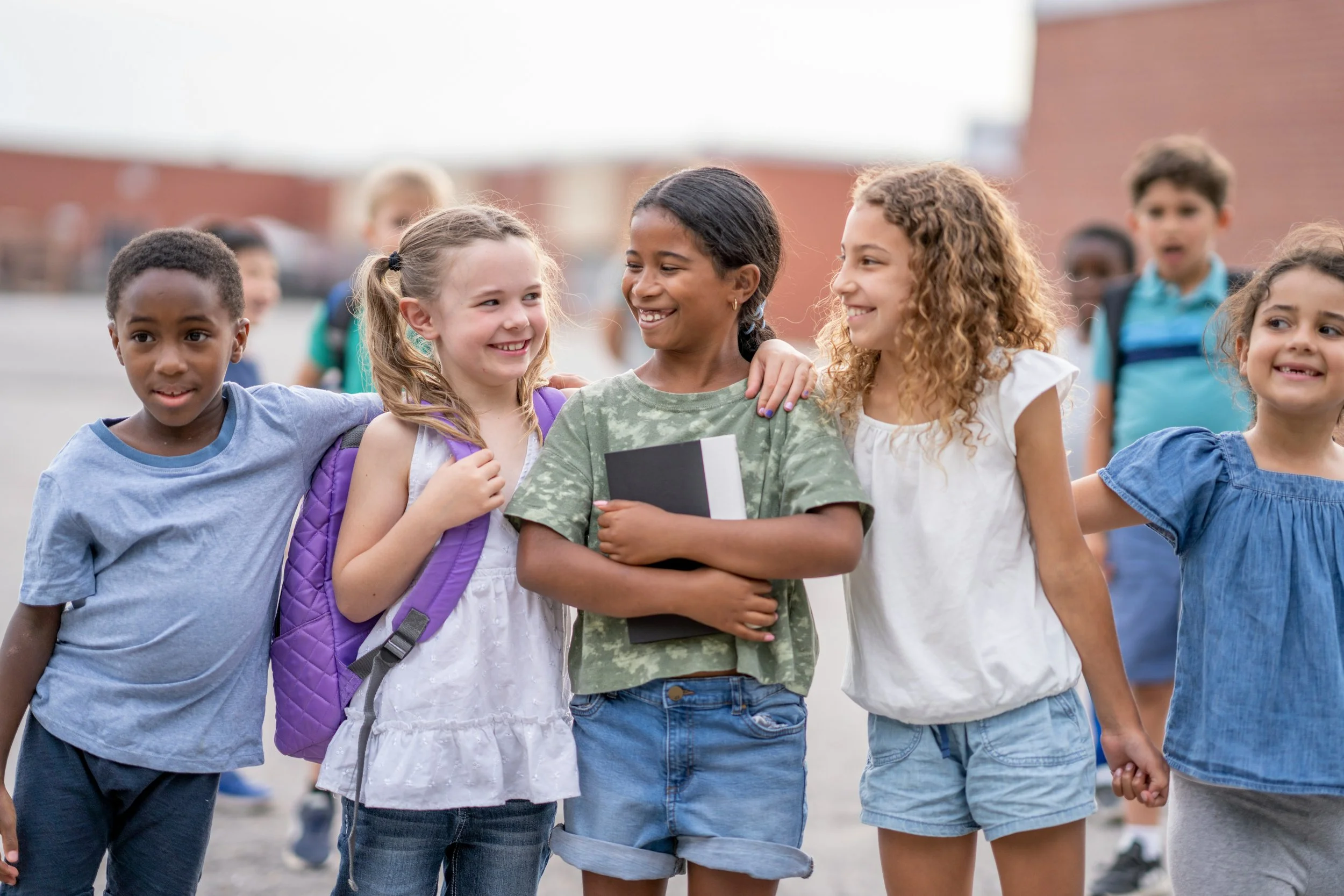 Group of diverse schoolchildren standing outside, smiling and holding hands with one girl in the center holding a book.