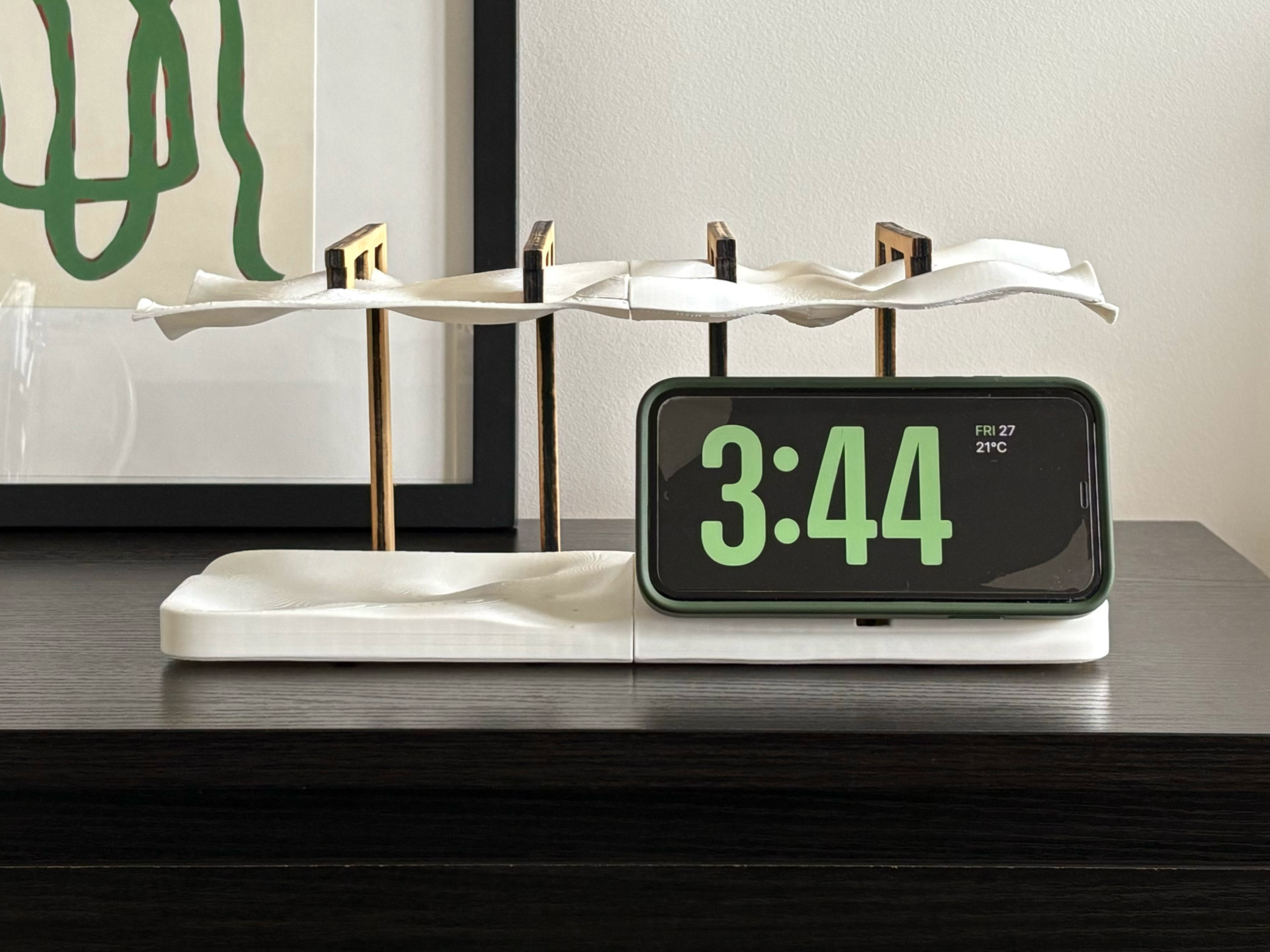 A modern clock showing the time 3:44, placed next to a decorative object with multiple vertical sticks and a white cloth on top, on a dark wooden surface.