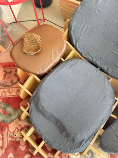 Close-up of two blue upholstered chairs with a wooden frame, positioned next to each other on a patterned red and green rug. Part of a brown circular stool with a woven piece resting on top is visible to the left.