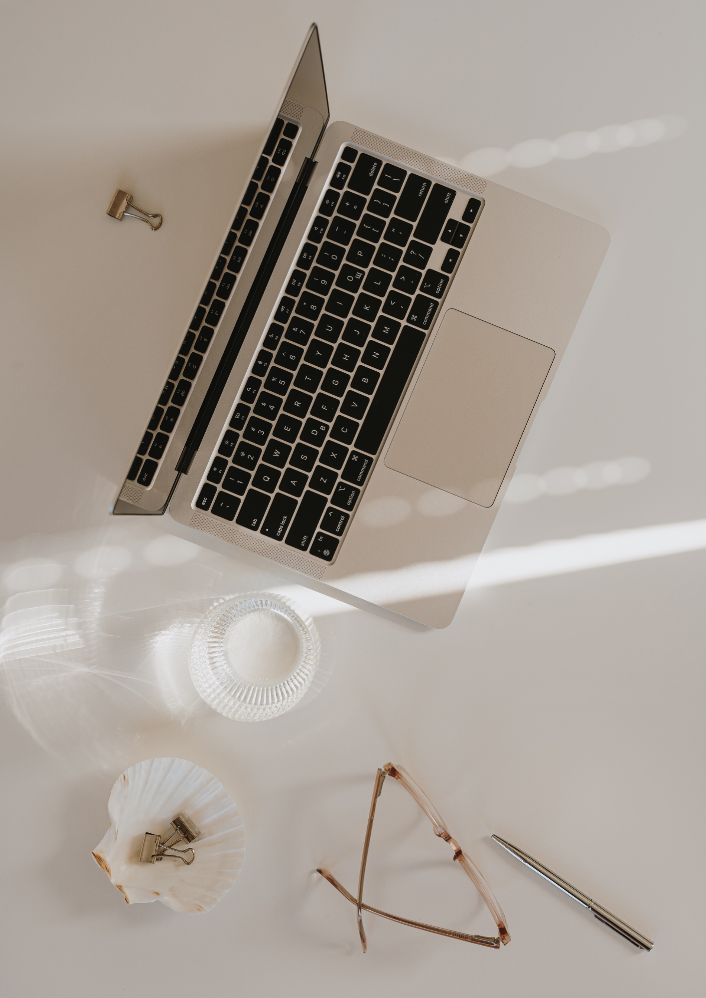 Top view of a silver laptop, a pair of eyeglasses, a pen, a seashell with binder clips, and a glass dish on a white surface.