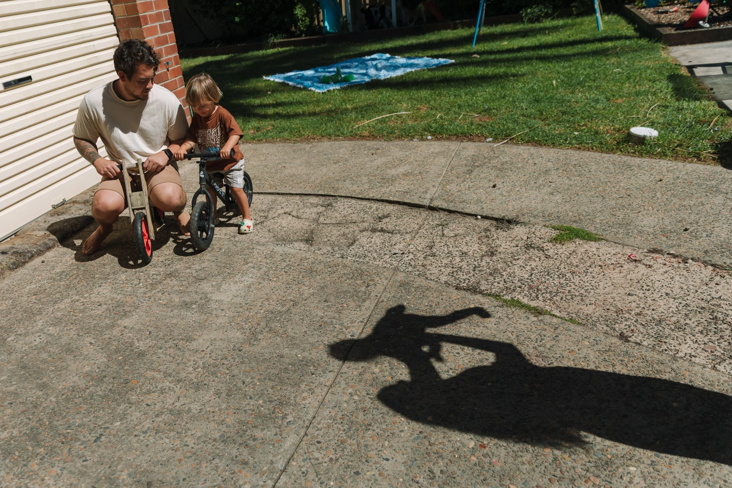 A man and a young boy are outside on a concrete patio, with the man helping the boy with a small bicycle. The man is kneeling and holding the bike, while the boy is walking beside it, holding the handlebars. There is a grassy area with a blue tarp in
