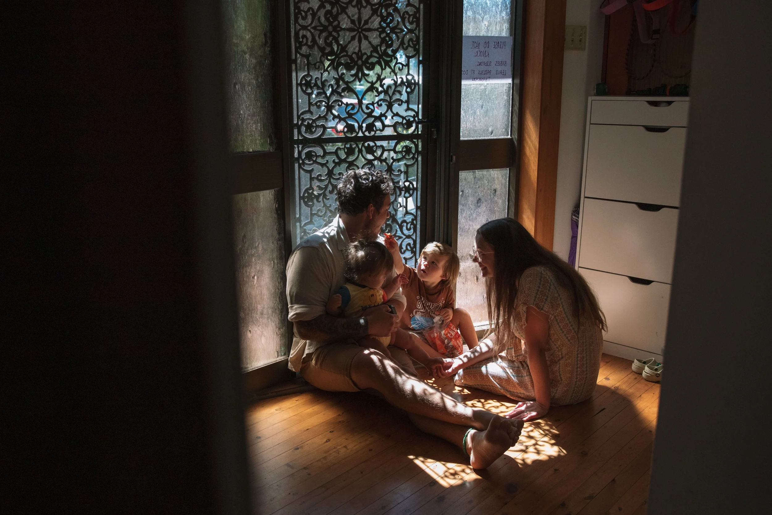 A family of four sitting on a wooden floor near a glass door, with sunlight shining through, engaged in a conversation. The father holds a baby, the mother and older child are looking at each other, smiling.
