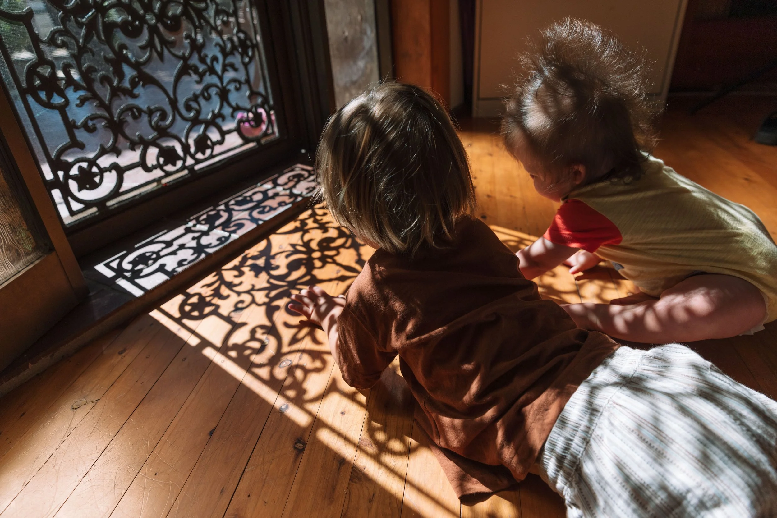 Two children sitting on a wooden floor, playing with shadows cast by sunlight through an ornate metal gate.