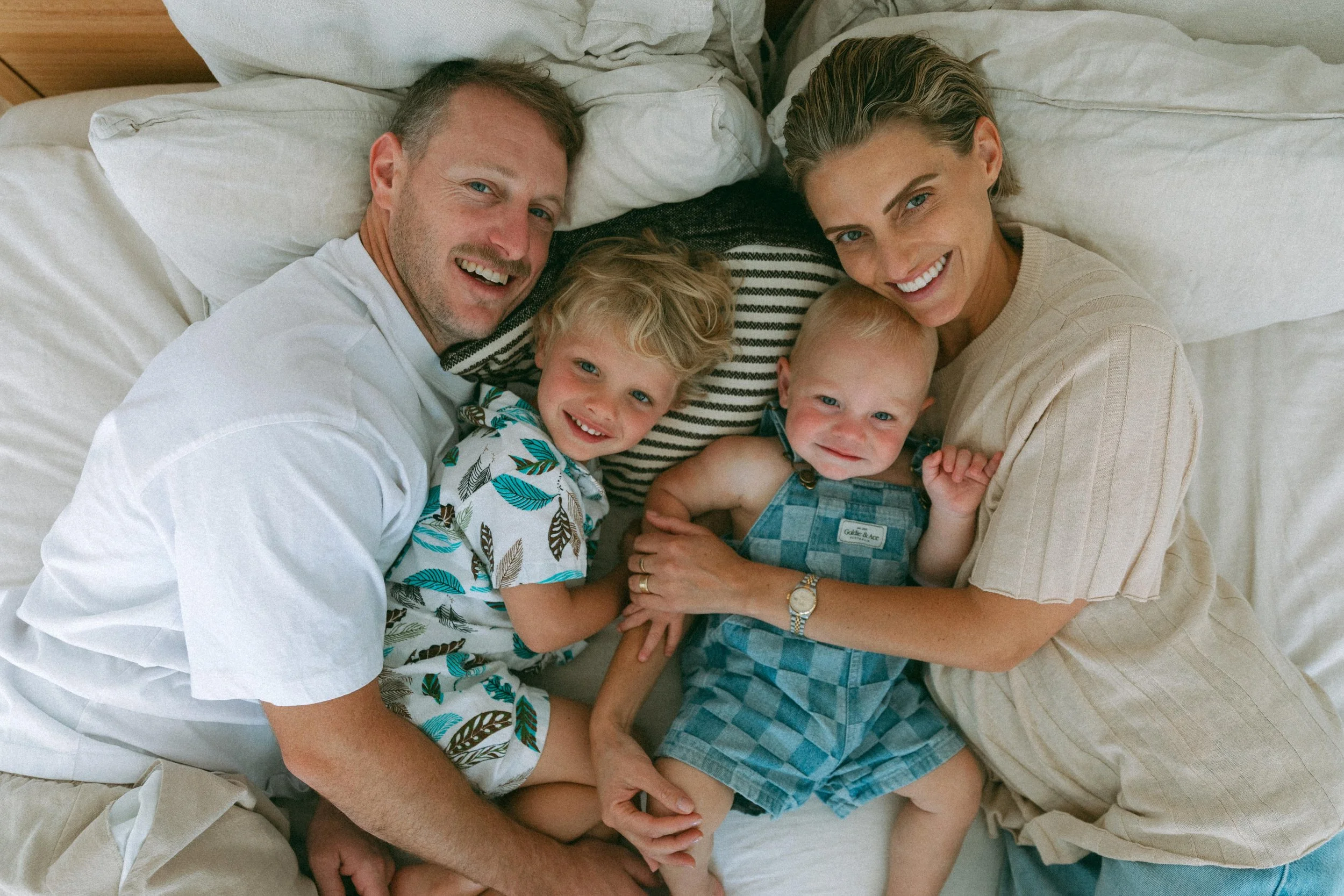 A smiling family of four lying on a bed, including a father, mother, and two young children, all looking up at the camera.