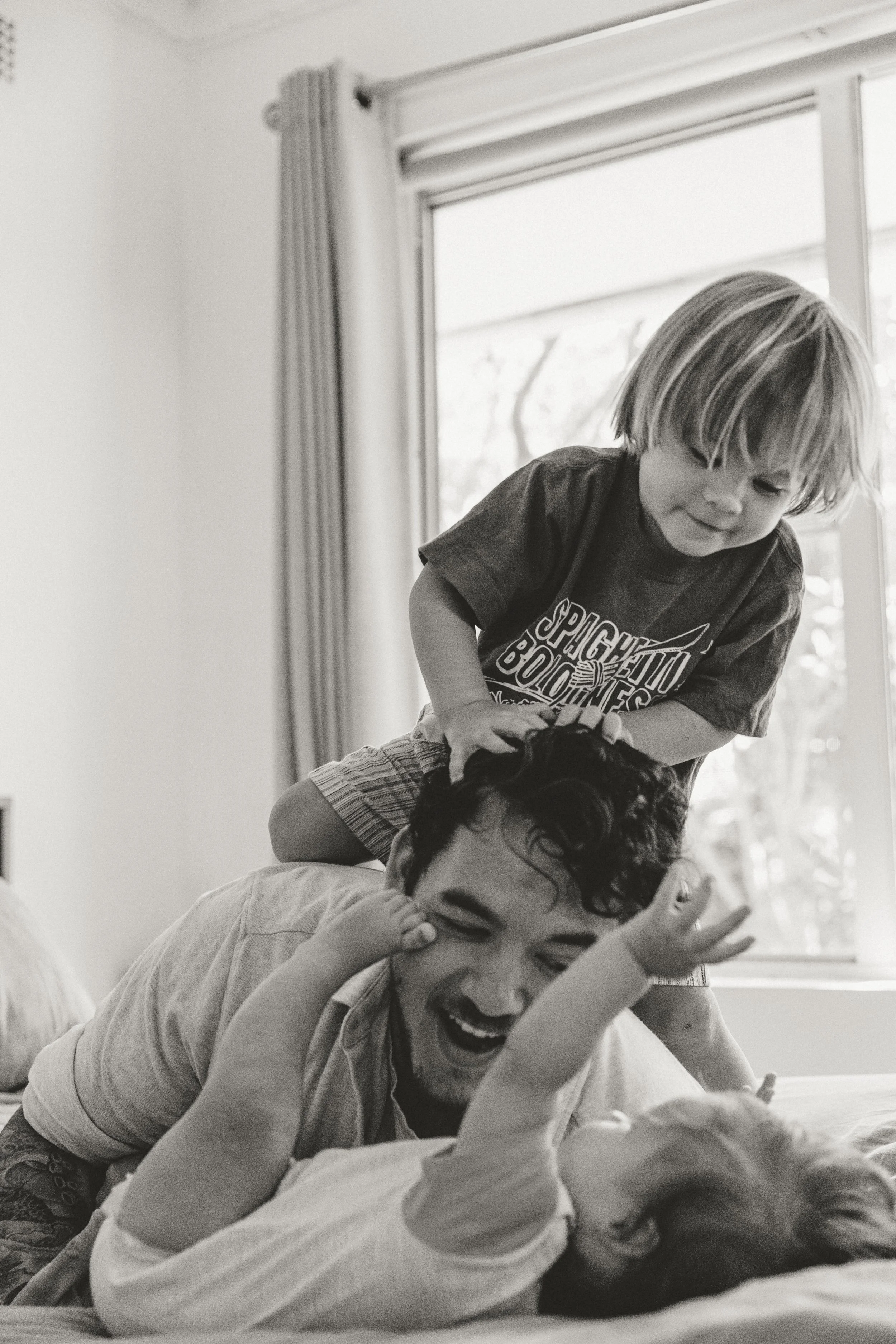 A joyful moment showing a man, woman, and child playing on a bed. The child is on top of the man, creating a playful scene indoors near a window.