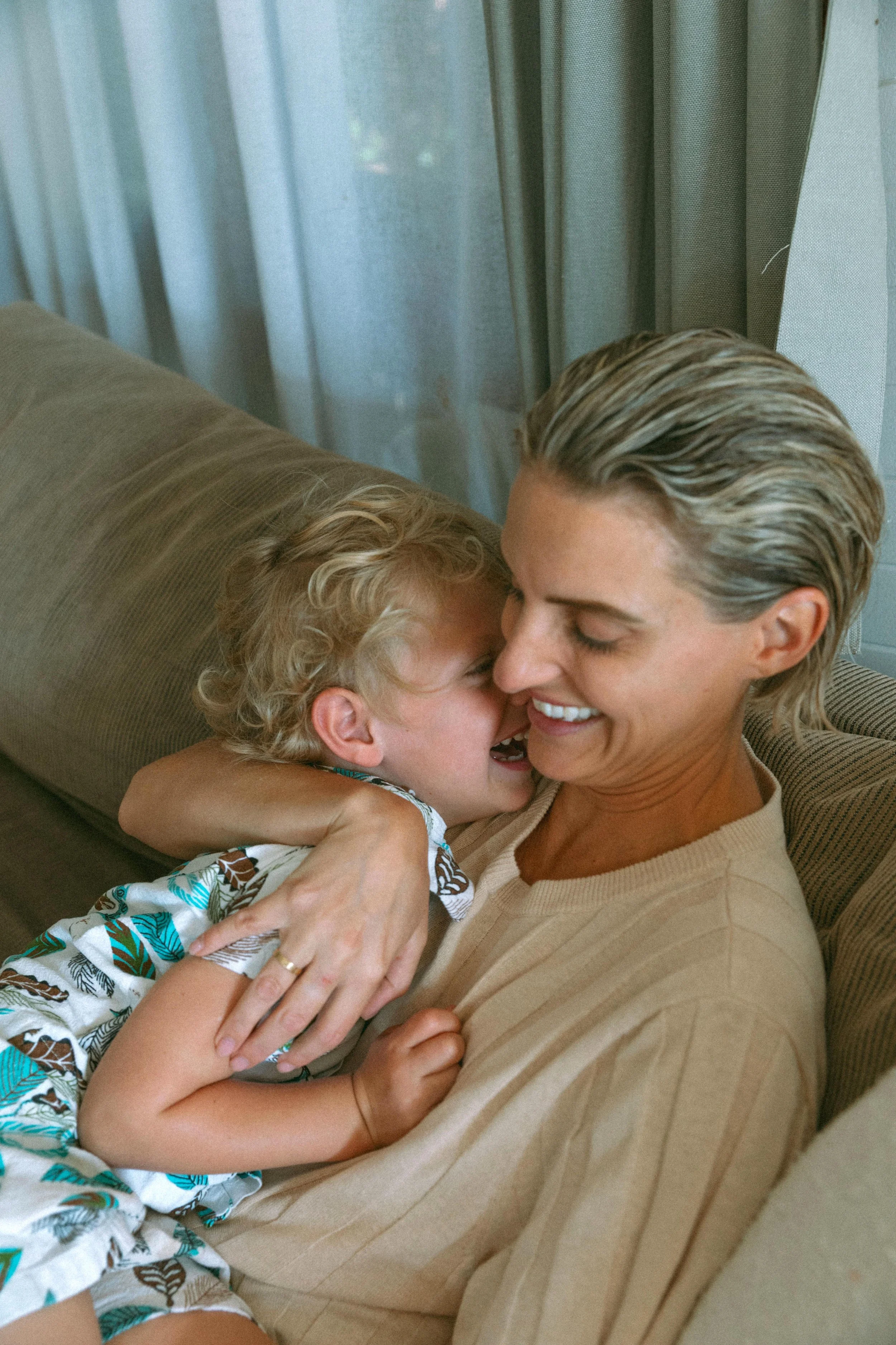 A smiling woman and a young boy sharing a joyful hug on a couch, with curtains in the background.