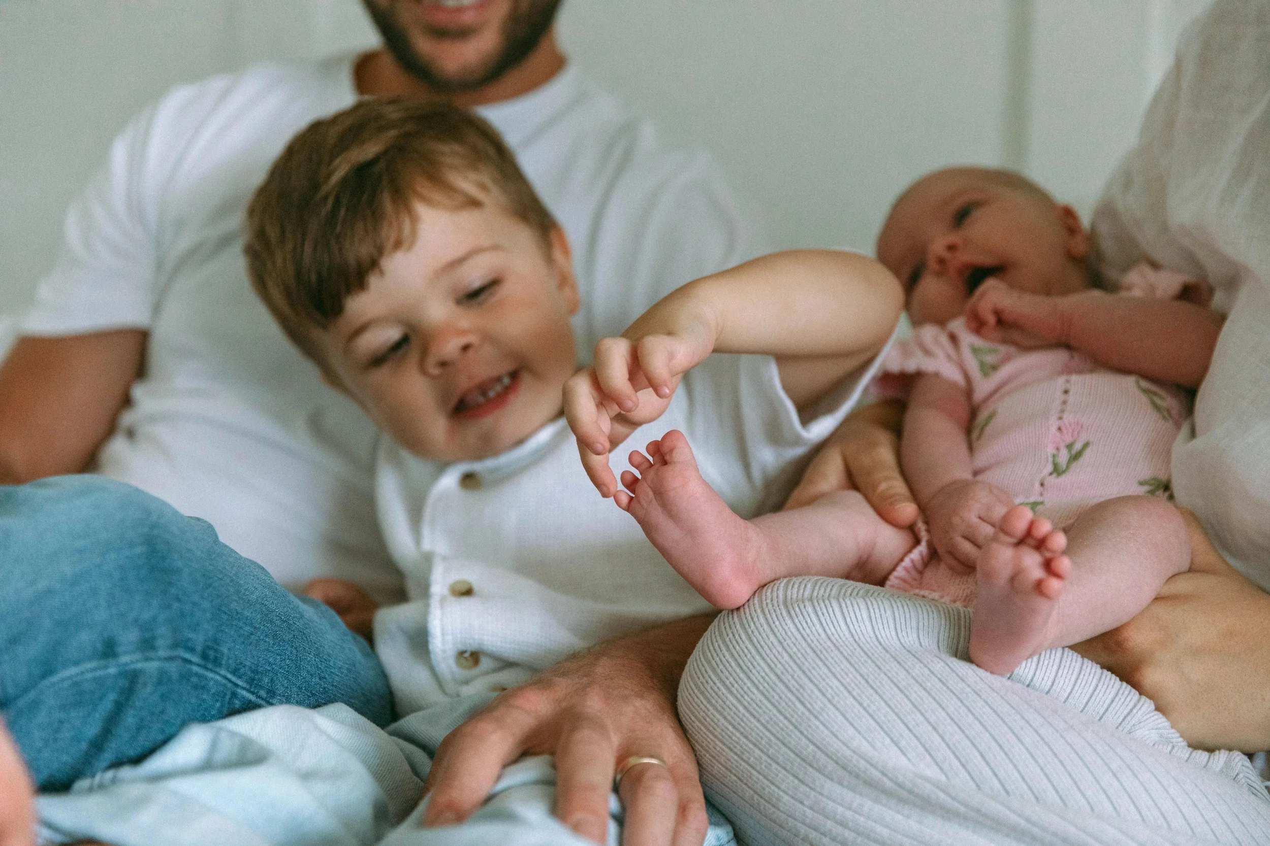 A young boy and a baby are lying on a bed with adults. The boy is reaching out to touch the baby's tiny foot, and both are smiling.
