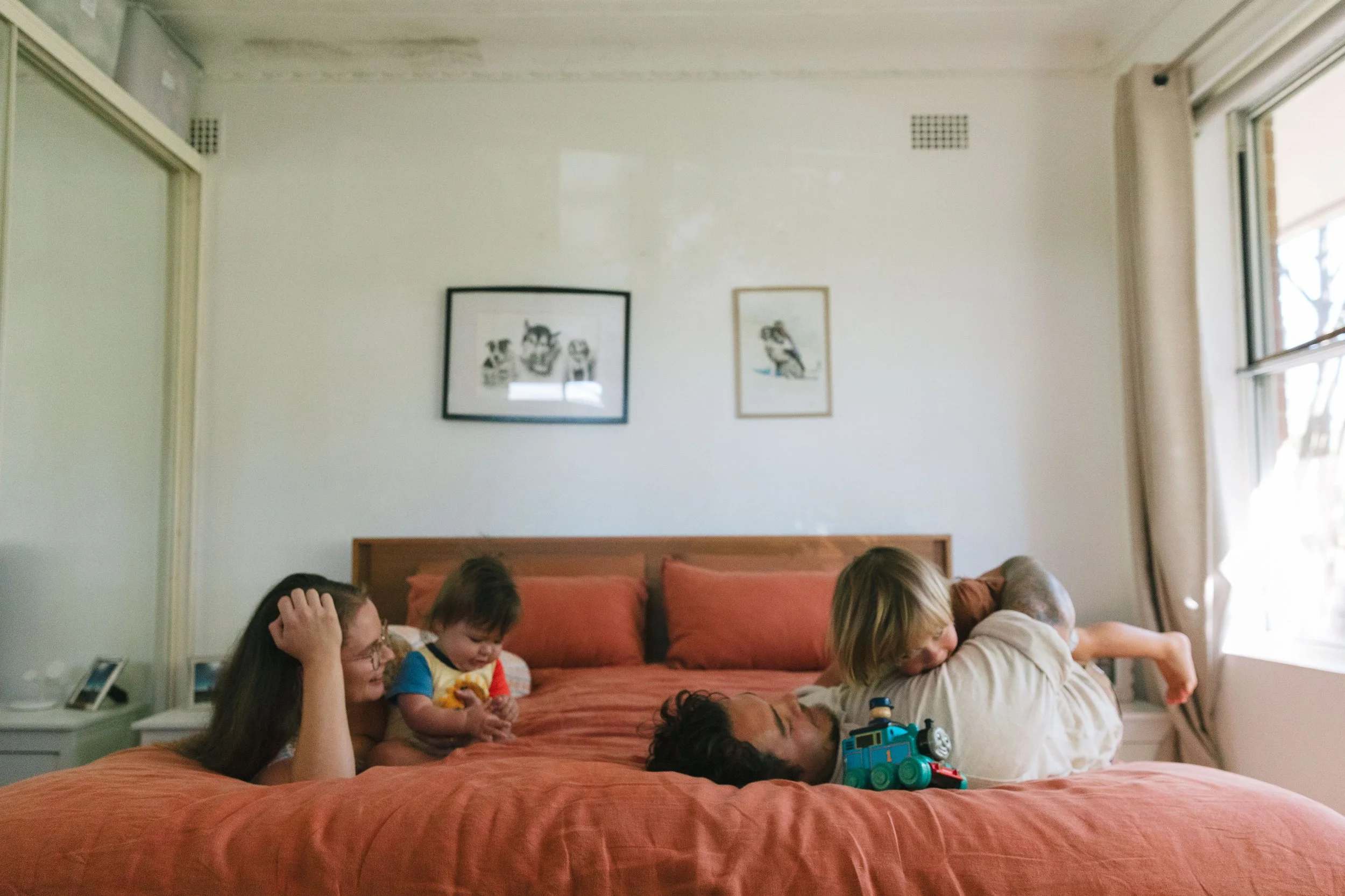 A family of five lying on a large bed in a bedroom, playing and cuddling. There are two children, a woman, and a man. The wall behind them is decorated with two framed pictures, and natural light is coming through a window with beige curtains.