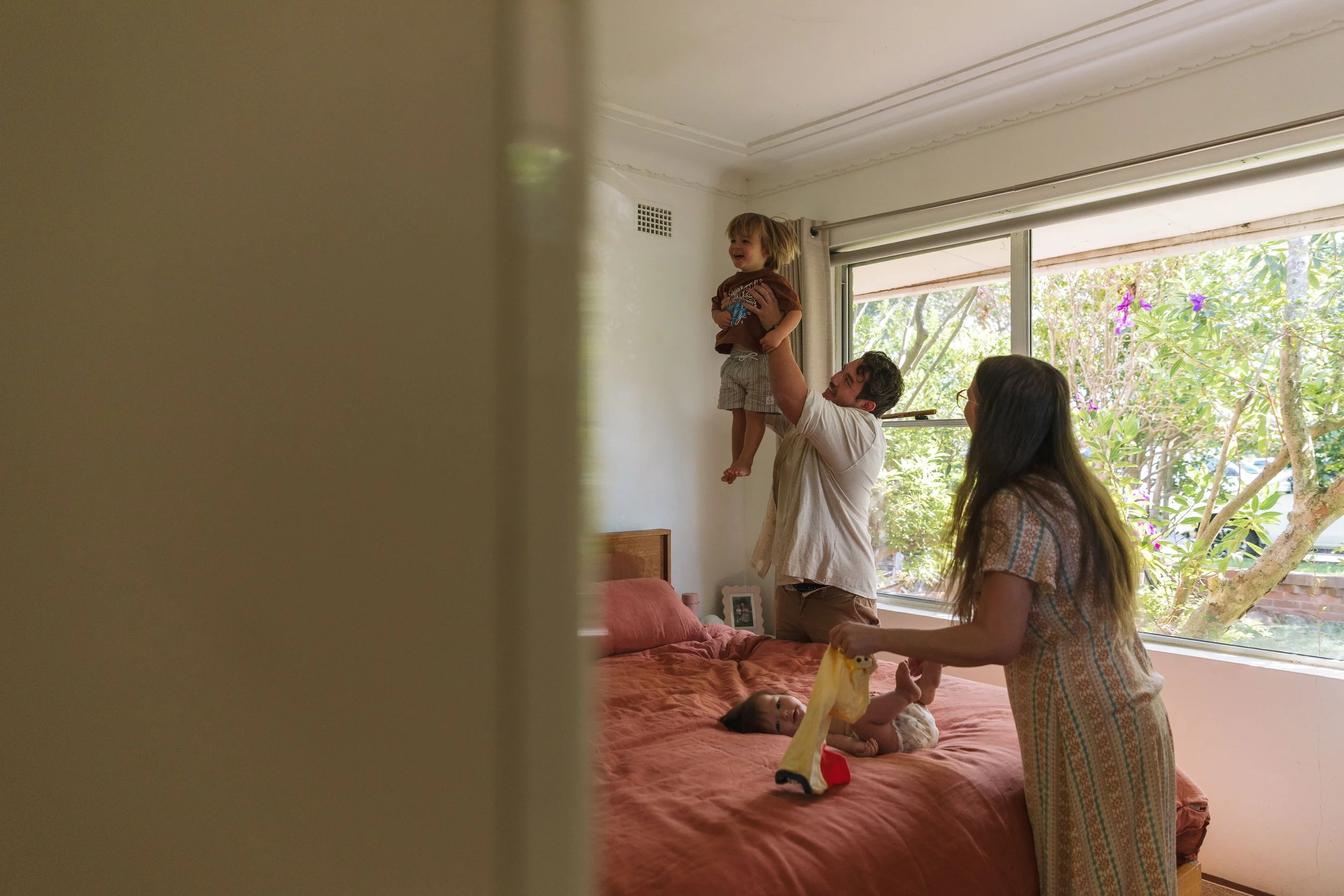 A man lifts a young boy in a bedroom, with a woman holding a cloth and a baby on the bed, all near a large window with trees outside.