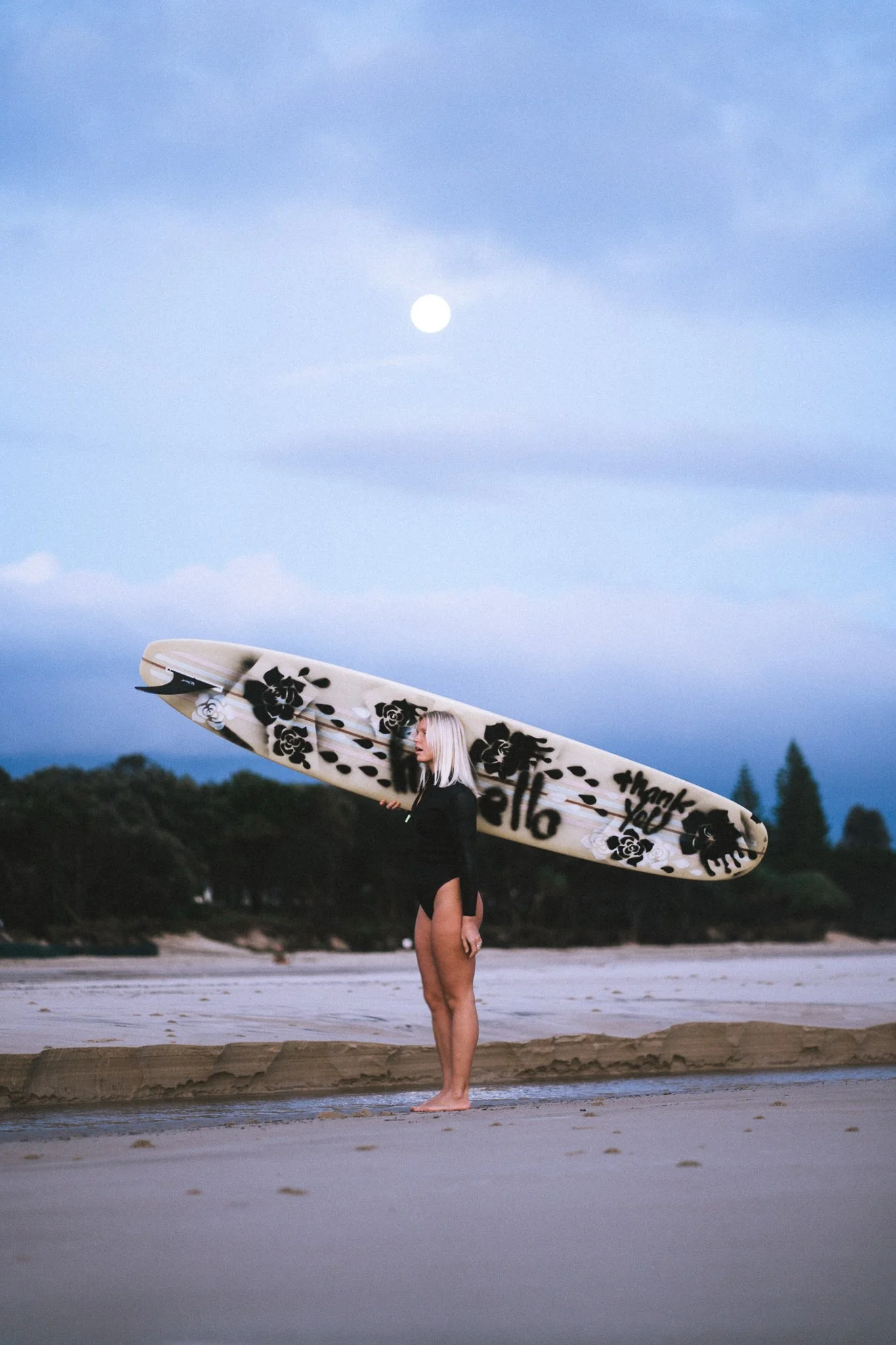 Sonja walking with her surfboard in Kintana wetsuit, Byron Bay headland in background.