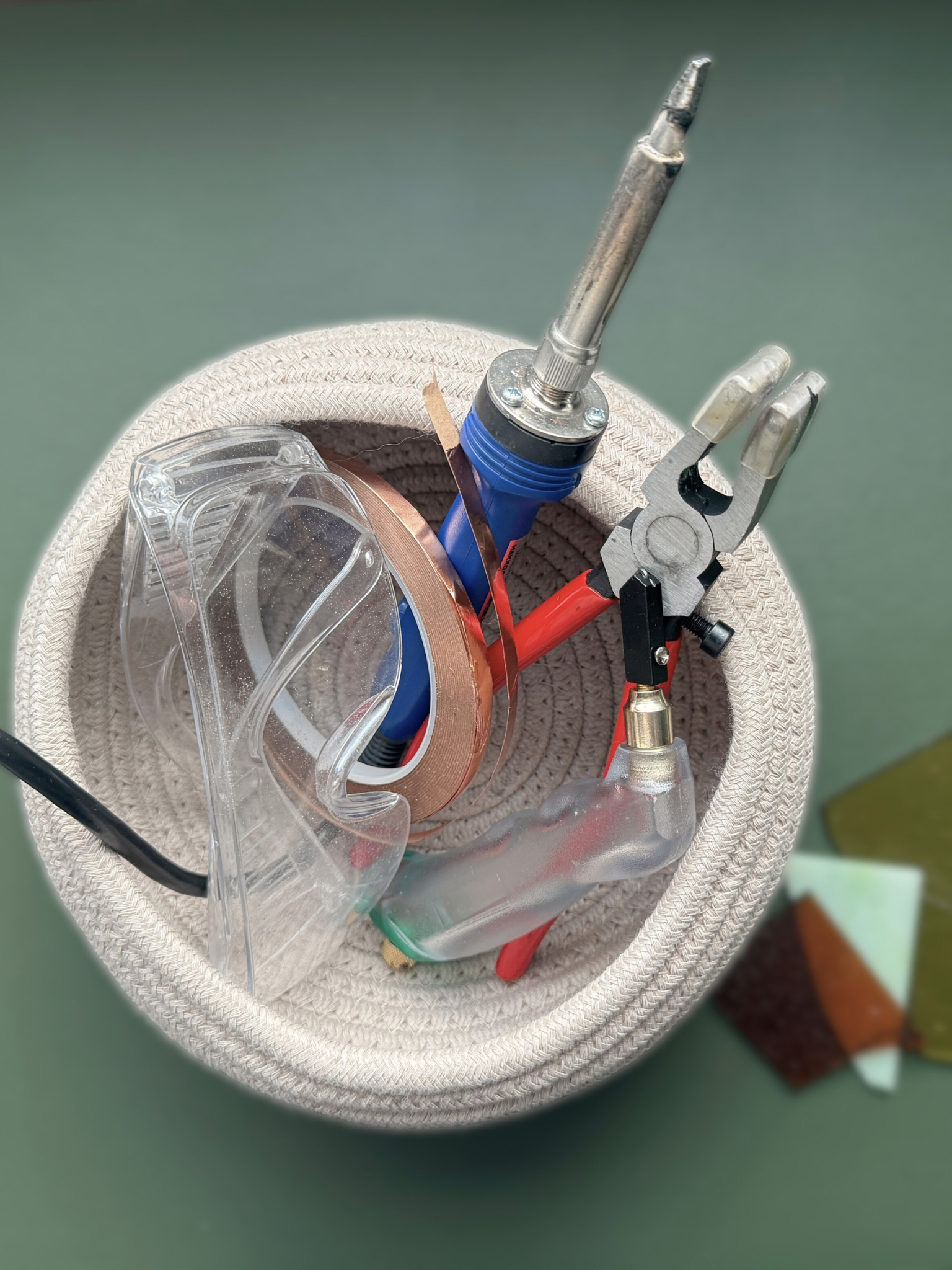 A cloth bucket containing various soldering tools including a soldering iron, wire cutters, heat-resistant gloves, and various colored sheets of abrasive material.