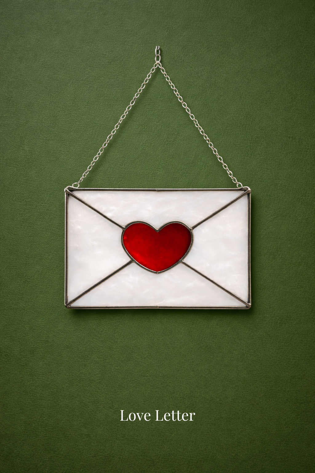 A stained glass decoration in the shape of an envelope with a red heart in the center, hanging on a green background. The text 'Love Letter' is written below.
