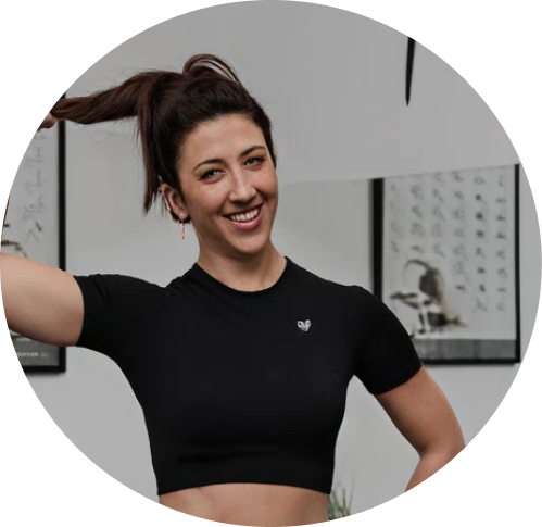 A woman with long brown hair wearing a black crop top smiling and touching her head in an indoor setting with artwork and a plant in the background.