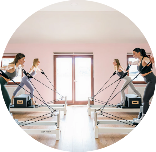 Four women exercising on Pilates reformer machines in a bright studio with a pink wall and a large window.