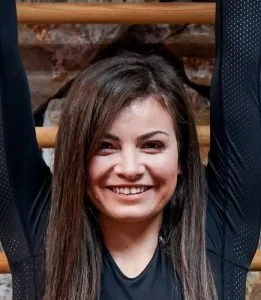 Young woman with long brown hair smiling, wearing a dark top, standing outdoors.
