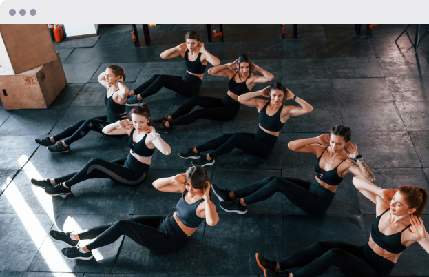 Six women in black workout outfits participating in an exercise class on a gym floor, sitting on the ground and performing side crunches with hands behind heads.