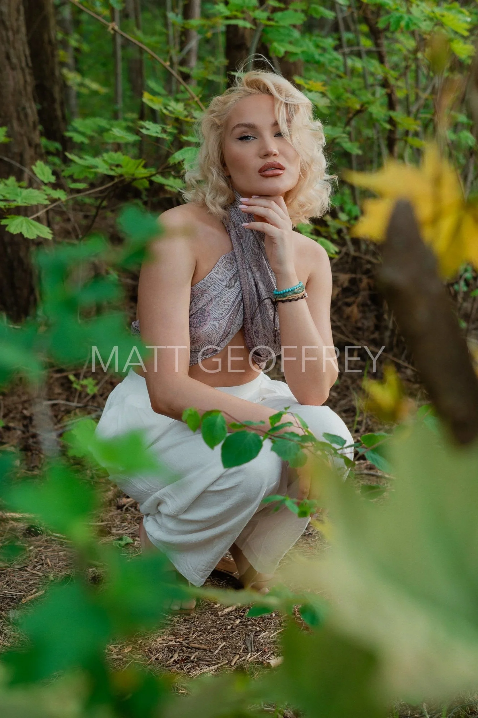 A woman with blonde curly hair and light skin crouches among green foliage in a forest setting. She wears a patterned halter top, white pants, and has bracelets on her wrist. She poses with her hand on her chin, looking confidently at the camera.
