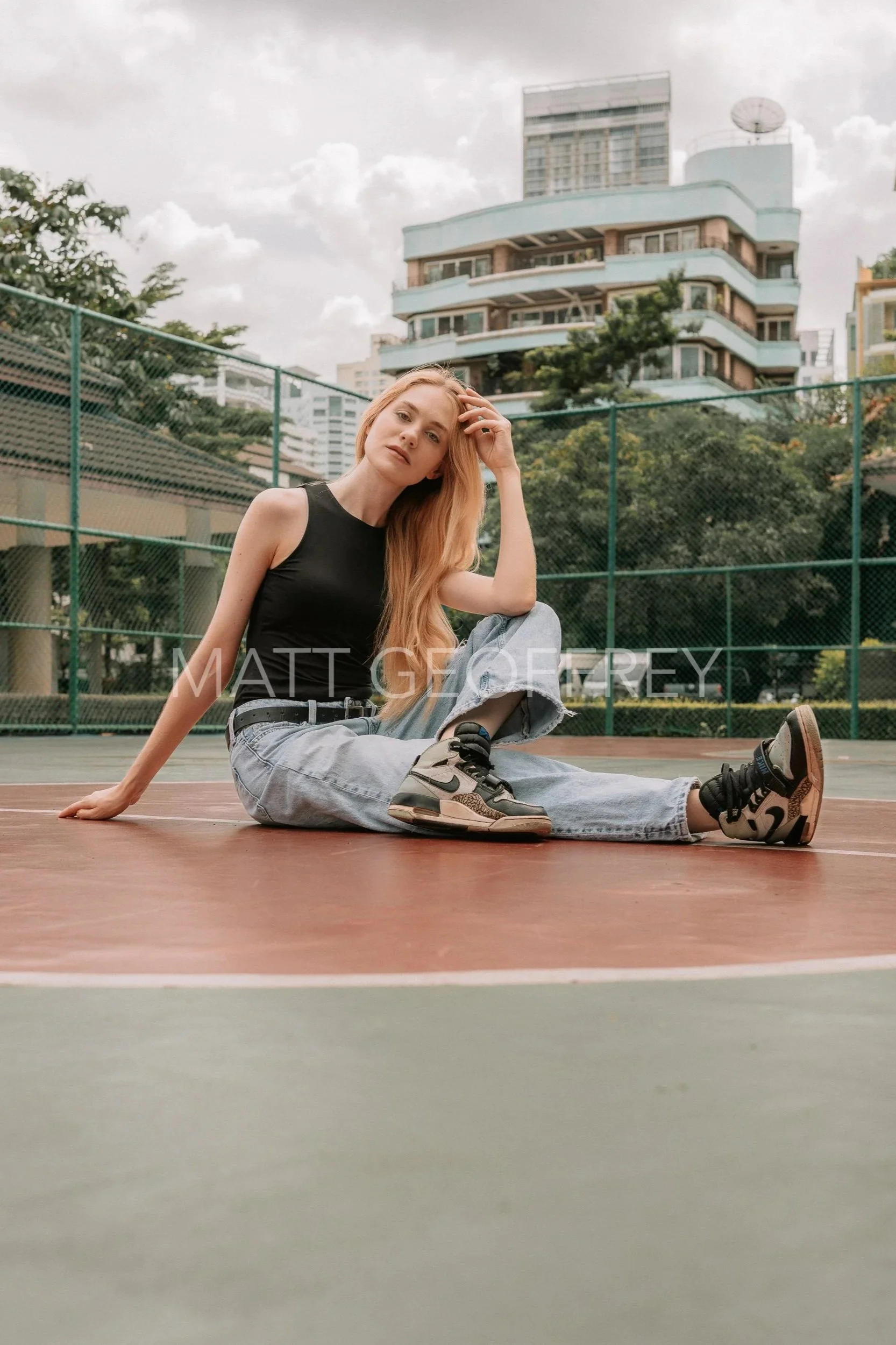 A young woman with long blonde hair wearing a black tank top, light blue ripped jeans, and sneakers sitting on an outdoor basketball court, with urban buildings and trees in the background.