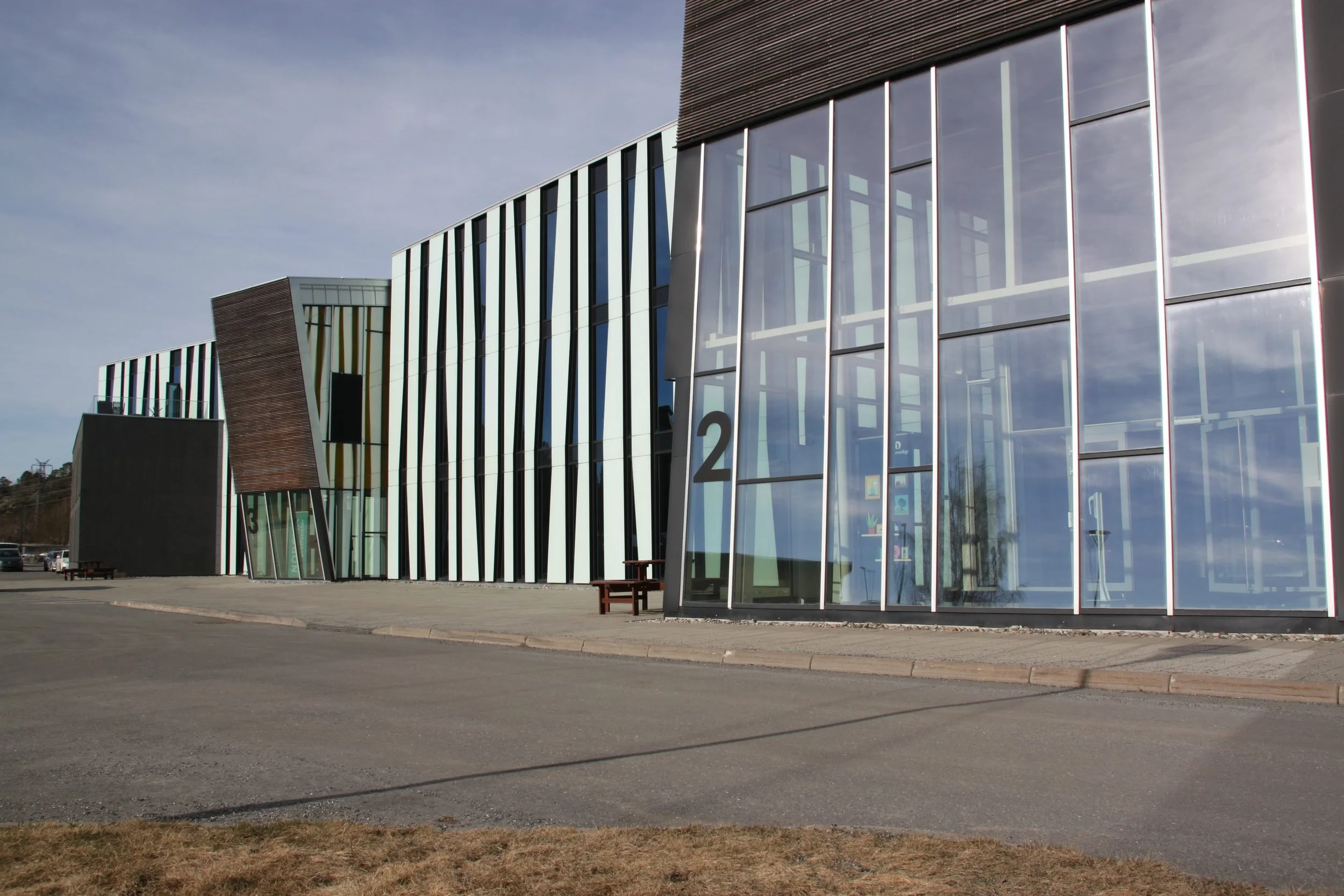 Moderne bygning med skrå glassfasader og svarte vertikale stripemønstre, med nummer 2 og 3 på veggen, omgitt av en veikant og parkeringsplasser.