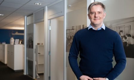 En mann står i en kontorbygning, smilende, iført blå genser.