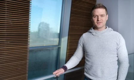 En mann står inne ved et vindu i et rom med tregulv og vegger. Han har på seg en lys grå genser.