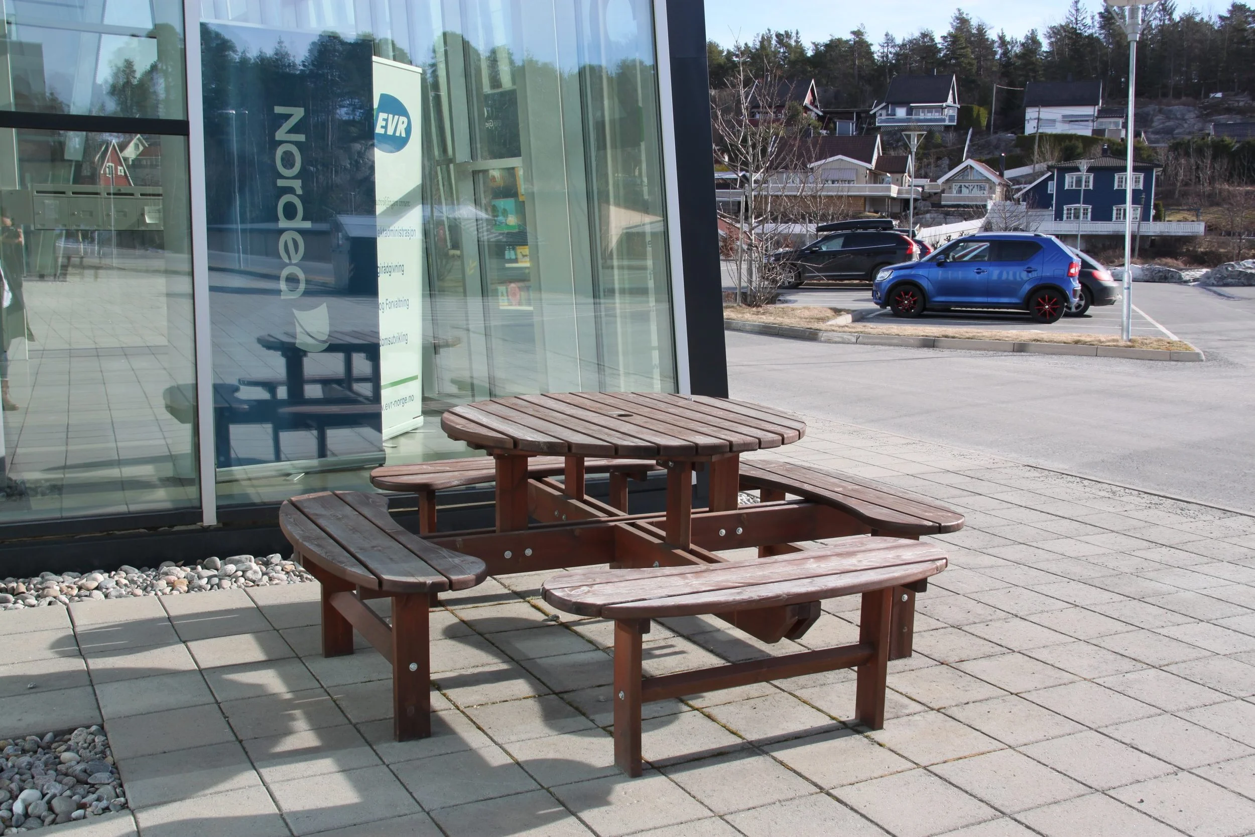 En utendørs benk og bord laget av tre plassert foran en glassfasade. I bakgrunnen er en parkeringsplass med flere biler og et boligområde med hus på en bakke. Det er sol og tydelig vær.