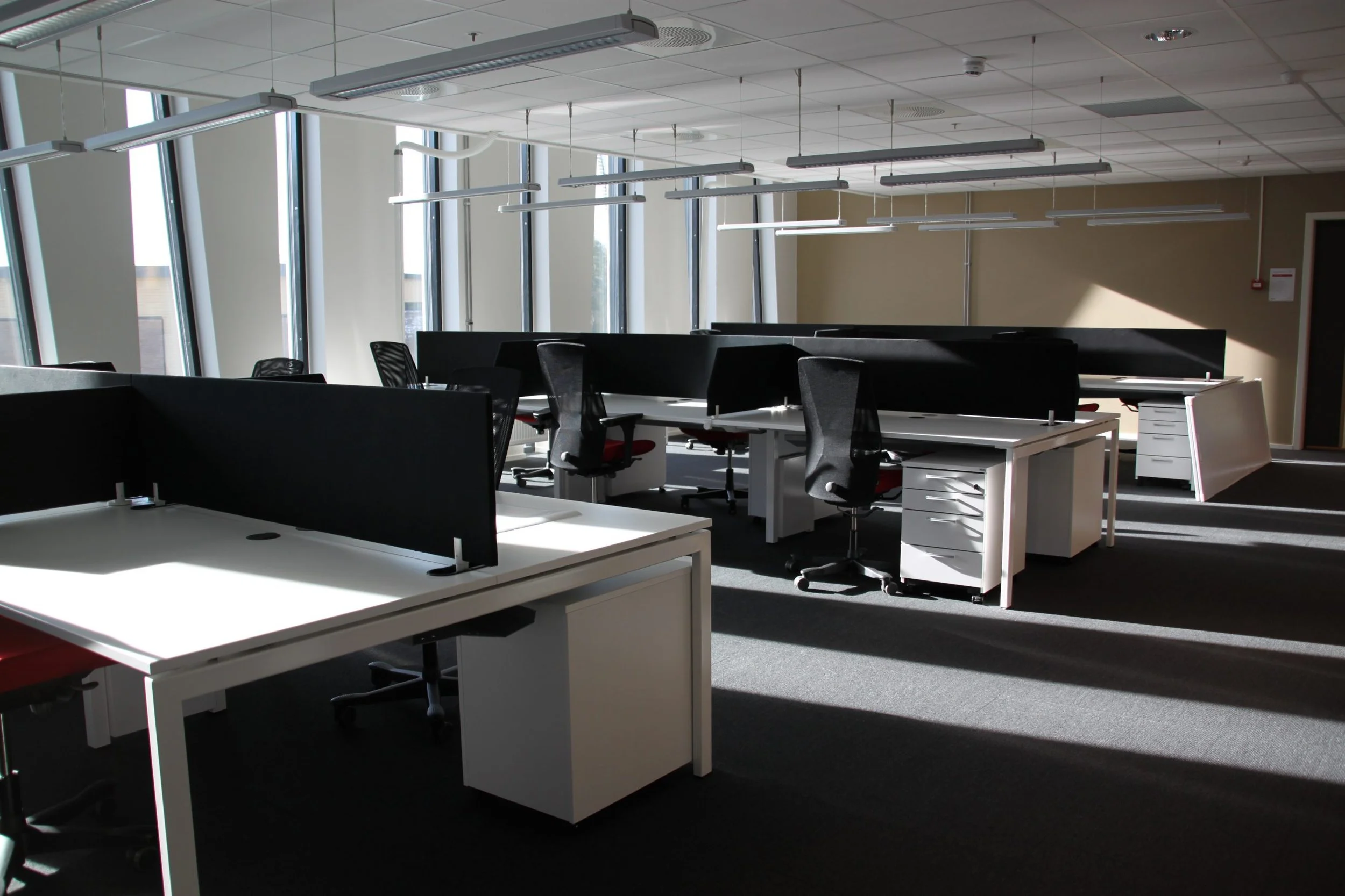 Kontor med flere arbeidsstasjoner, skrivebord og kontorstoler, store vinduer som slipper inn dagslys.